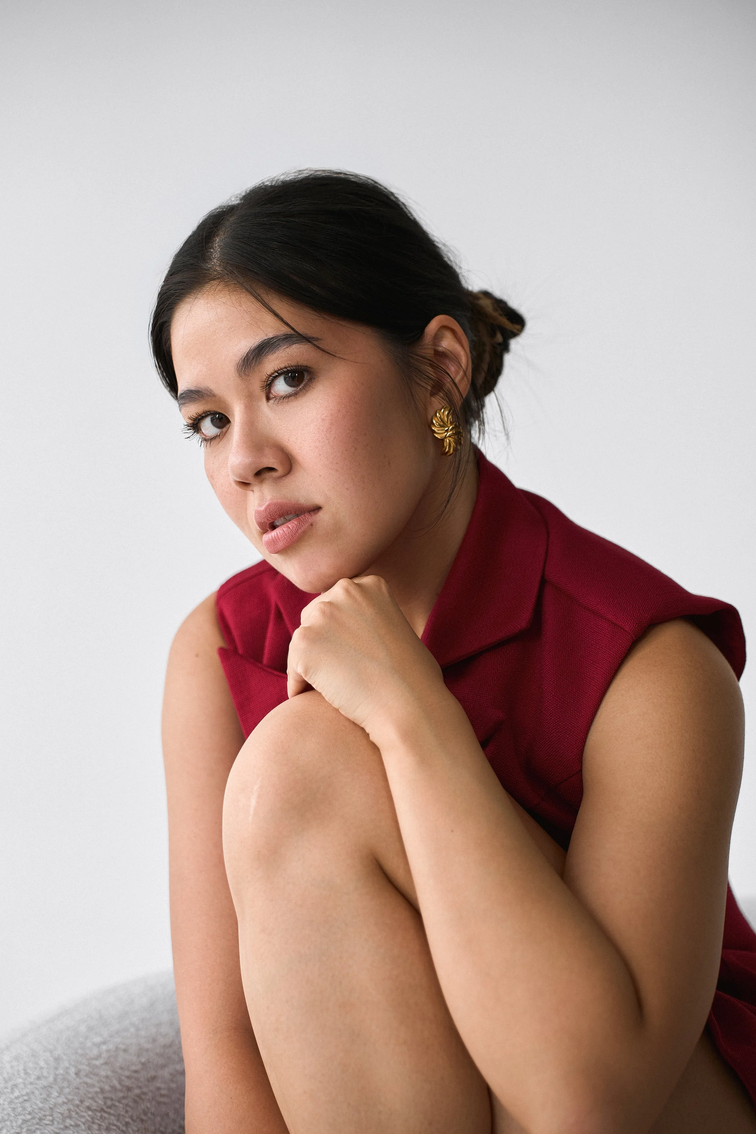 A woman with dark hair styled in a bun, wearing gold earrings and a sleeveless maroon top, resting her chin on her hand against a plain gray background.
