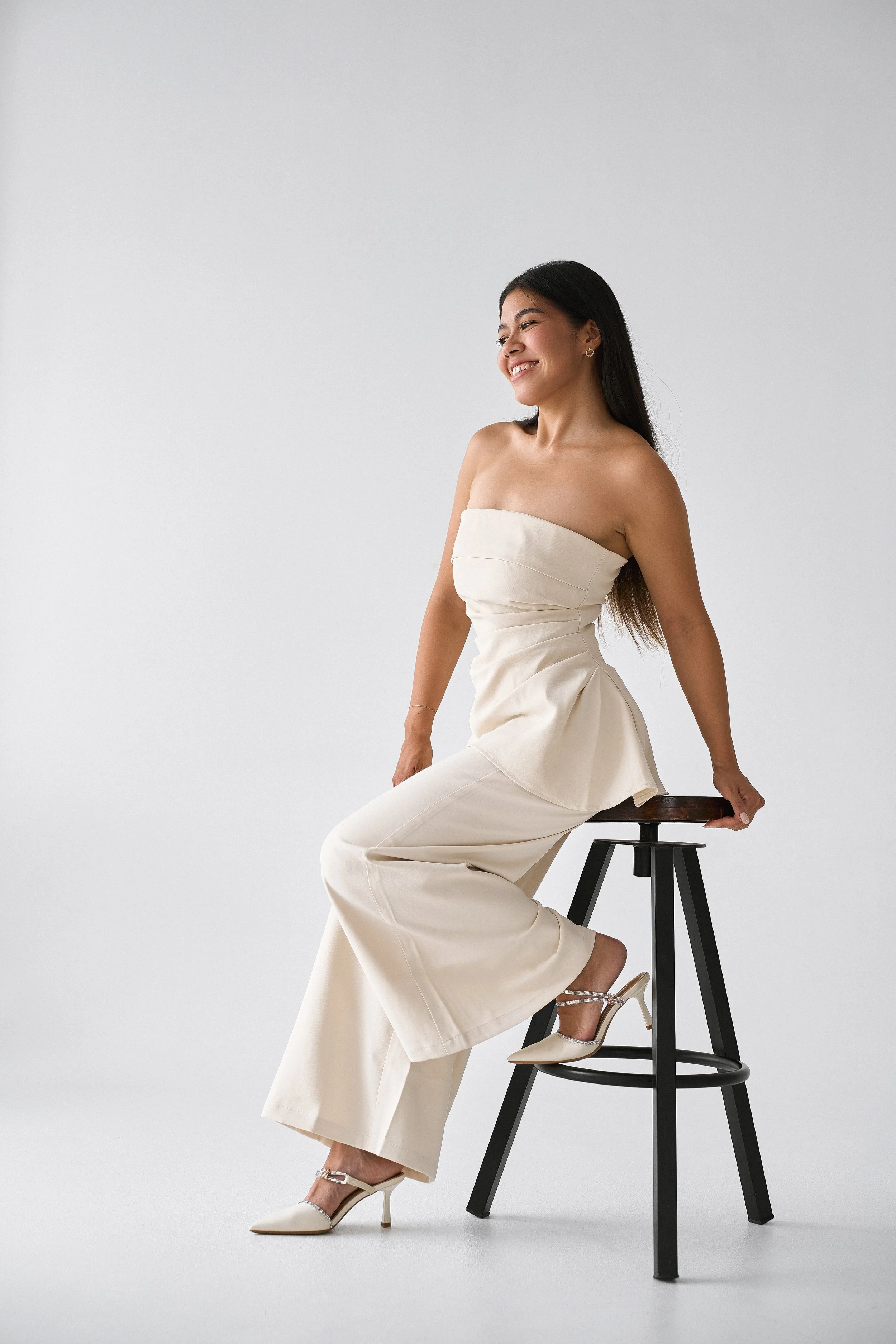 A young woman with long dark hair sits on a black wooden stool, smiling and looking to her left, wearing a strapless cream-colored dress and high heels, against a plain light gray background.