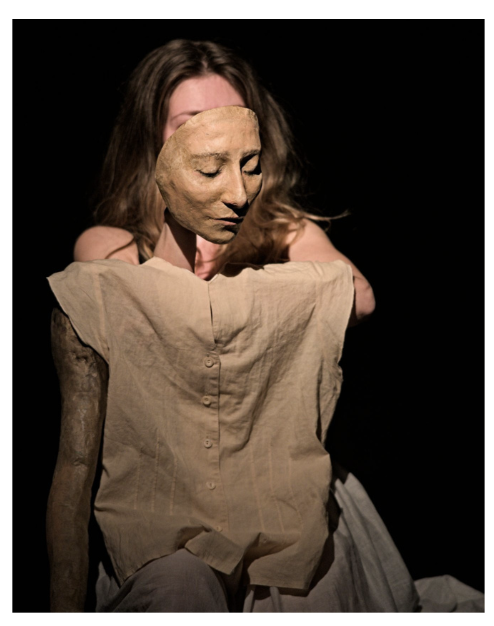 Una scena teatrale con una donna che tiene un manichino con un volto di cartapesta, evidenziando emozioni intense di tristezza o introspezione.
