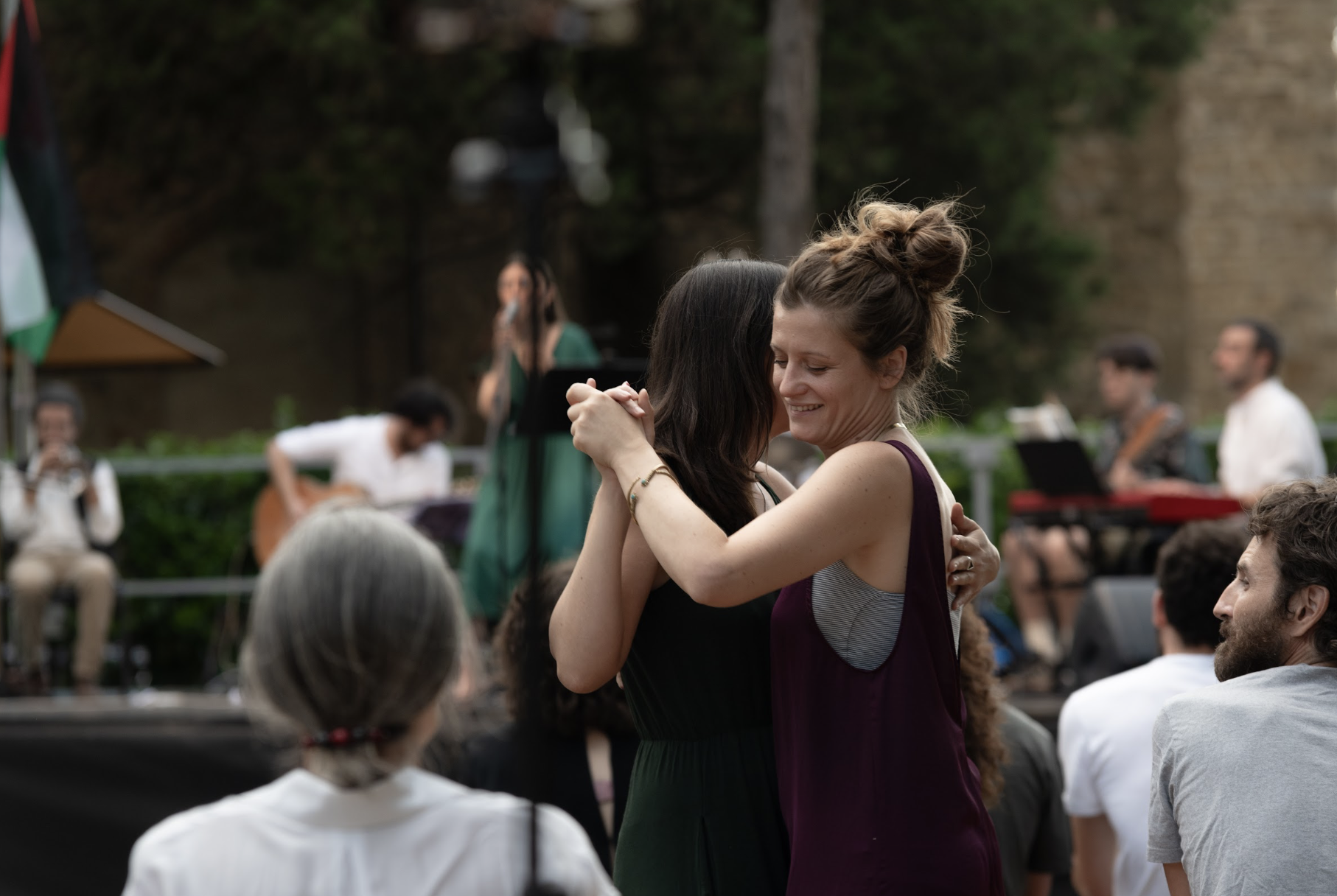 Due donne si abbracciano e ballano in un evento all'aperto con un pubblico seduto e musica dal vivo in sottofondo.