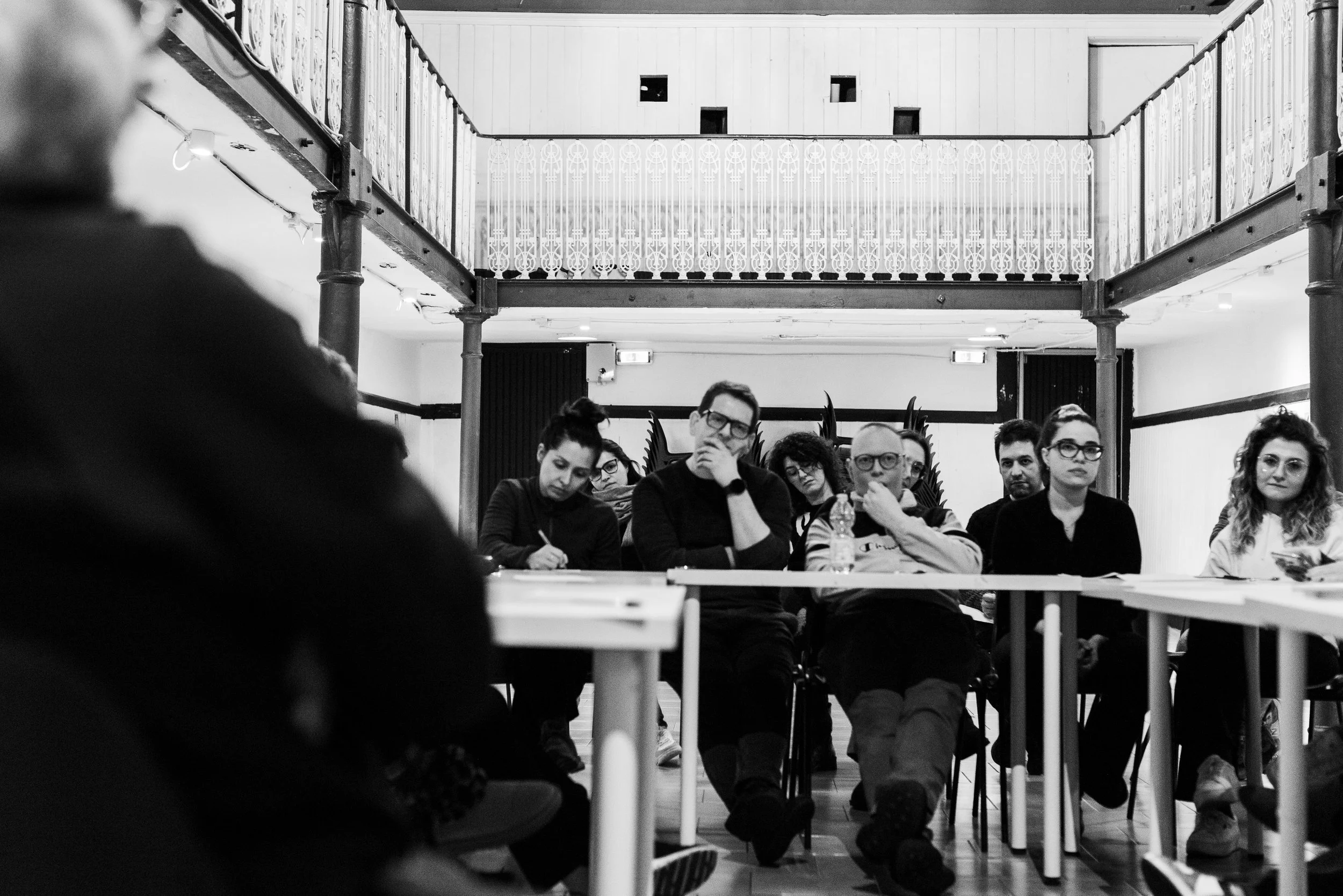 Un gruppo di persone siede a tavoli durante una riunione o una conferenza, in un ambiente interno con una balaustra decorativa sullo sfondo.