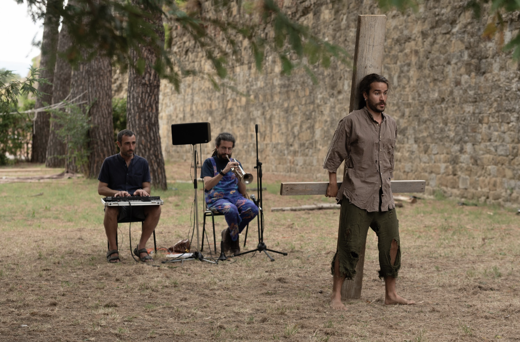 Un uomo con abiti logori e senza scarpe, legato a un palo, che rappresenta Gesù Cristo, un trio di musicisti che suona strumenti musicali, tra cui un sintetizzatore, una tromba, e un microfono, in un'area all'aperto con alberi e un muro di pietra sul