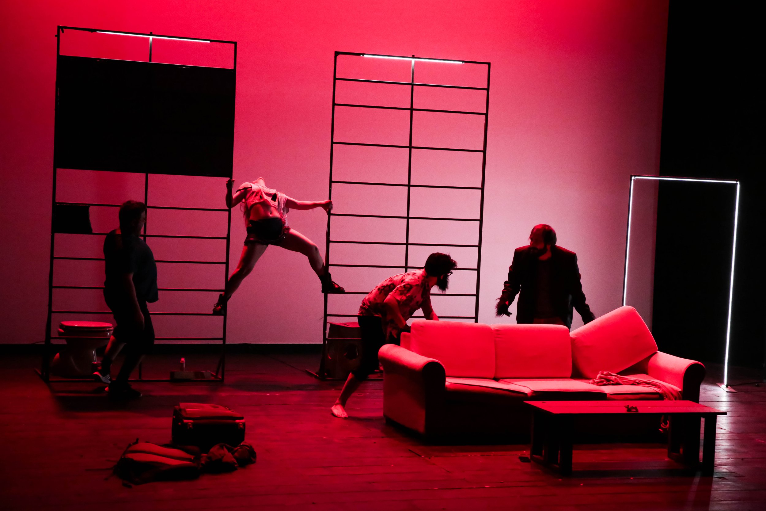 Scene teatrale con attori che si muovono su un palco rosso con elementi di scenografia minimalisti, tra cui divano rosso, scaffali e una piattaforma.