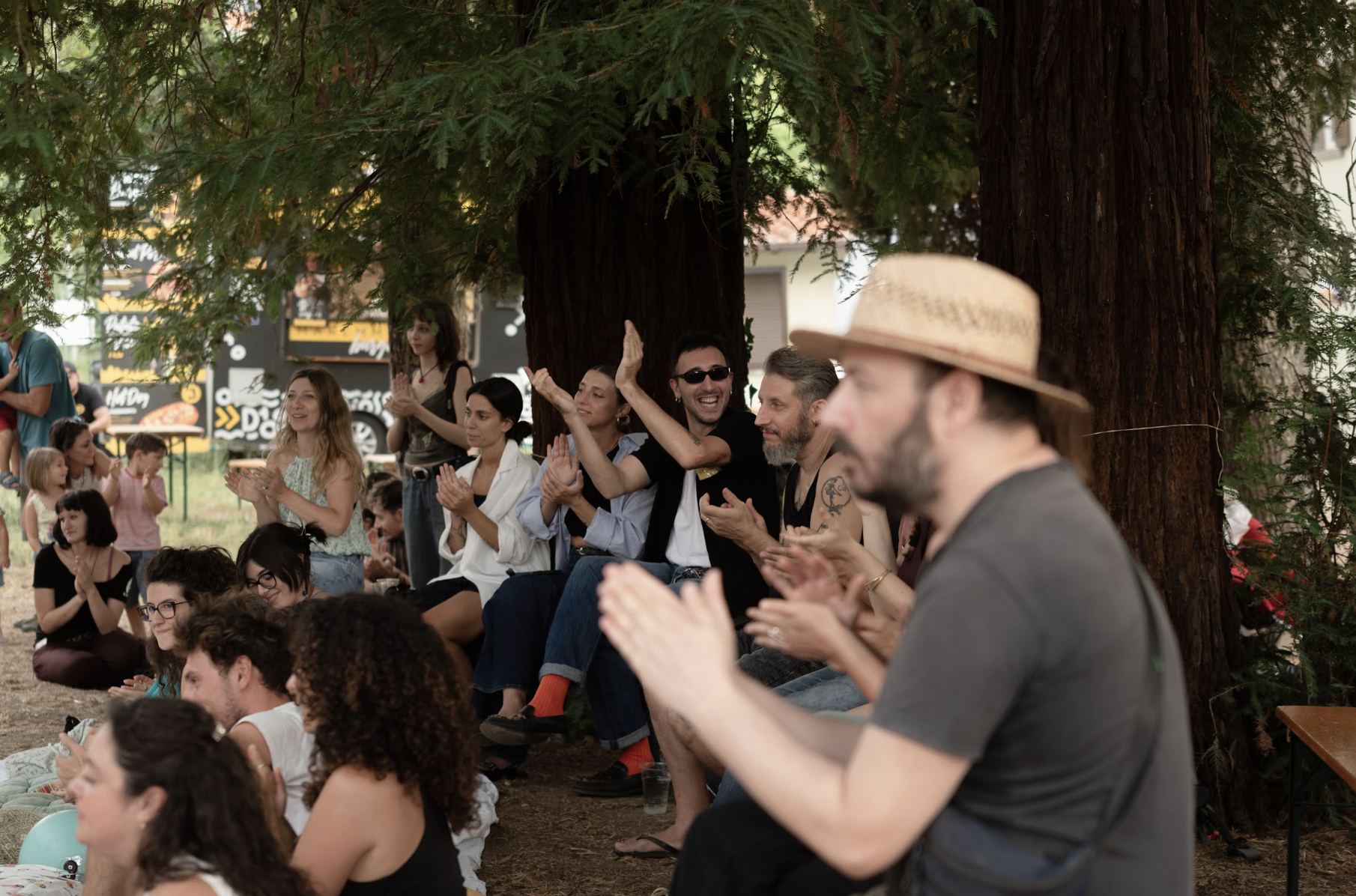 Un gruppo di persone sedute sotto grandi alberi, che applaudono e sorridono, in un ambiente aperto e naturale, durante un evento o una manifestazione.