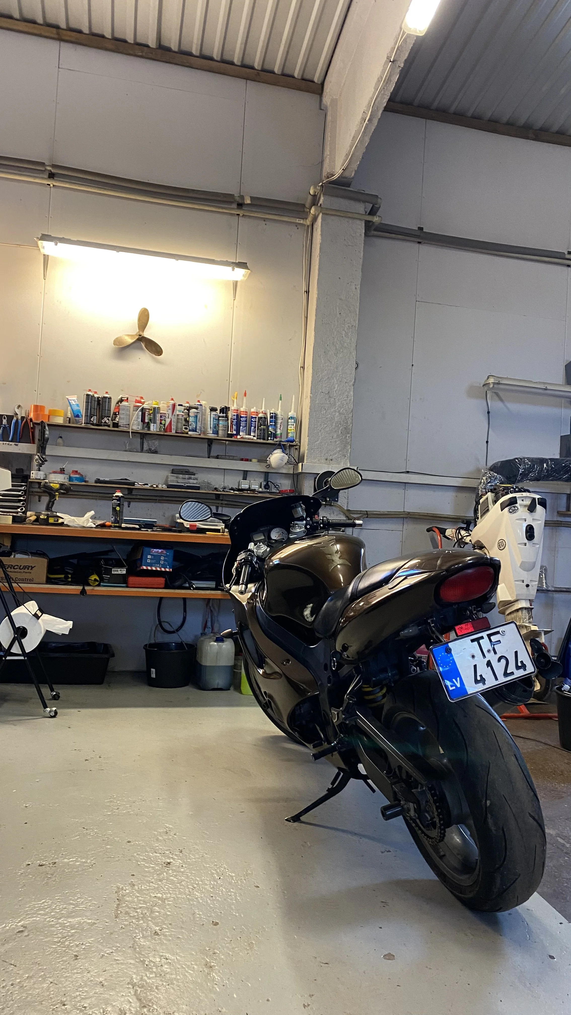 A motorcycle parked inside a garage. The garage has tools, supplies, and shelves in the background, with a wall-mounted fan and fluorescent lighting. The motorcycle has a license plate reading "TF 4124."