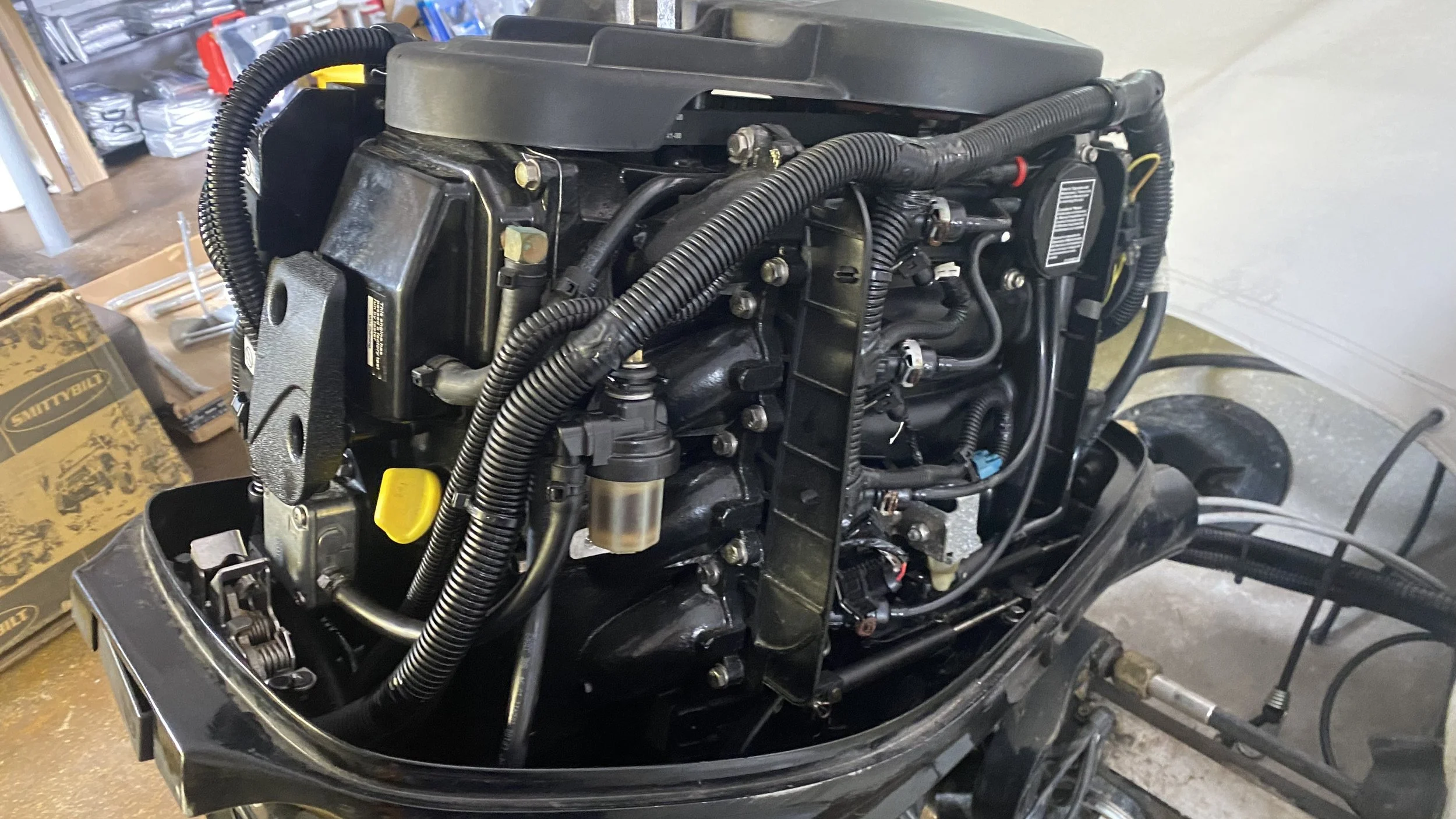 Close-up of an outboard motor with exposed engine components and wiring, placed on a boat in a workshop or garage.