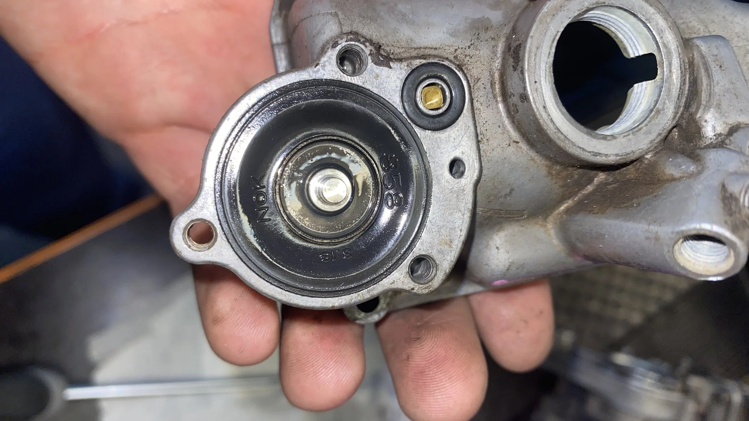 A close-up of a person's hand holding a metal automotive component with a black circular part and threaded holes.