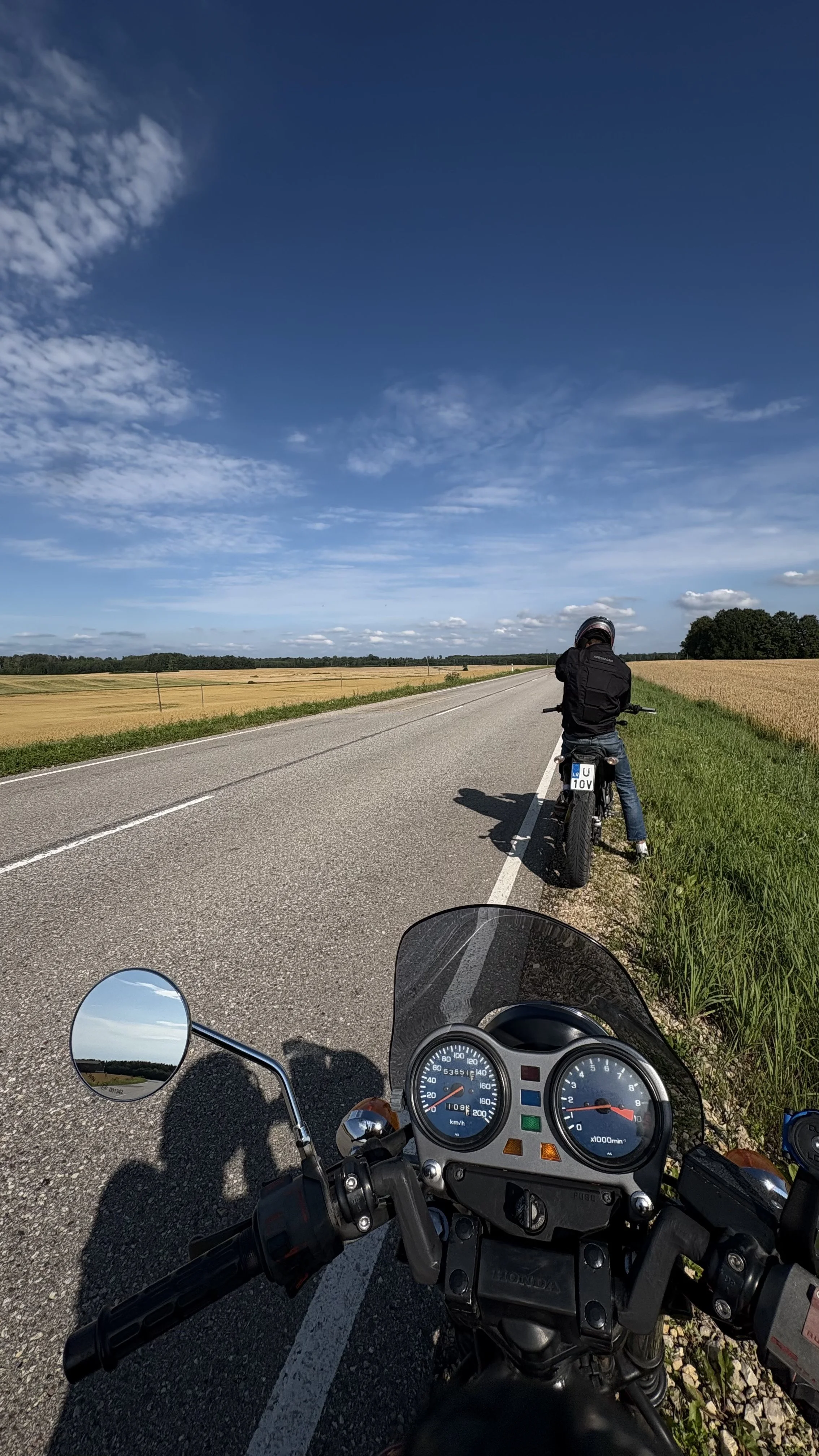 Motorcycle dashboard with gauges and a rider's view of a person wearing a helmet, sitting on a motorcycle on the side of a rural open road with fields and blue sky.