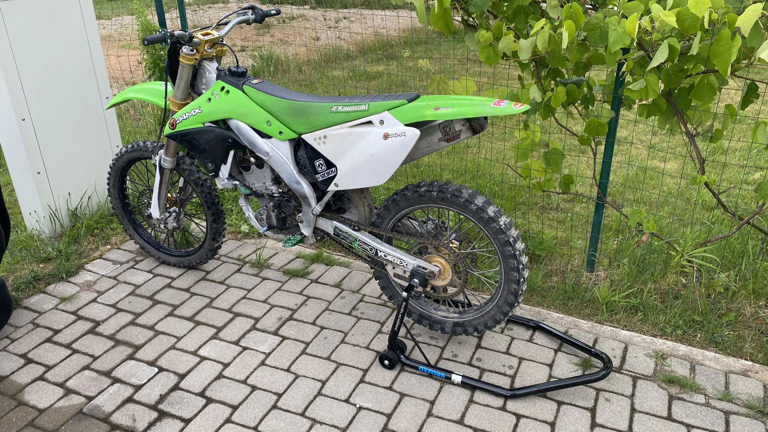 A green Kawasaki dirt bike resting on a padded stand on cobblestone pavement, with a background of grass, a green vine-covered fence, and a white utility box.