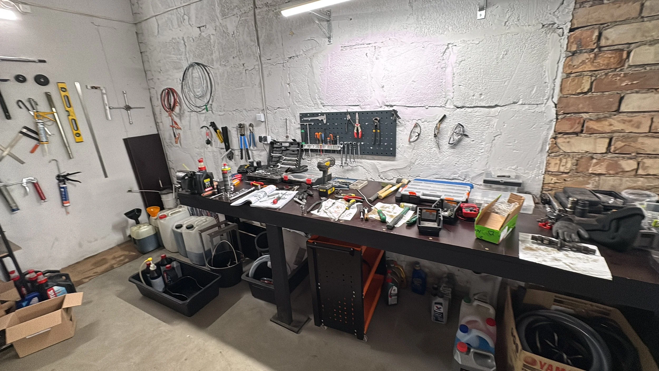 A cluttered workbench in a garage or workshop with various tools hanging on the wall and scattered on the table, including screwdrivers, pliers, a cordless drill, and containers of lubricants and sprays. There are also bins with supplies underneath t