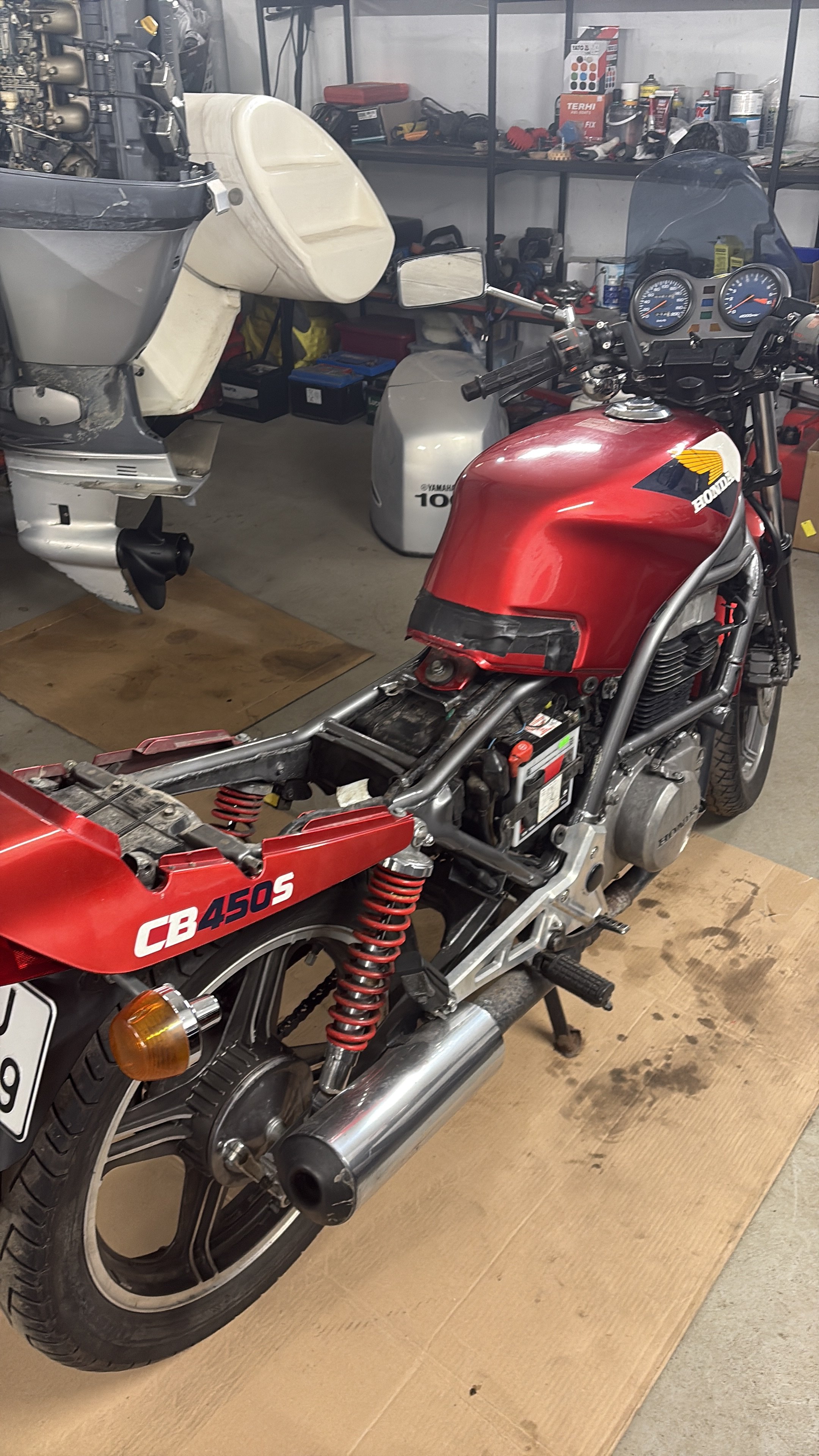 A red Honda motorcycle with part of the body removed, revealing the engine and suspension. A white outboard motor is mounted nearby, and shelves with tools and supplies are visible in the background in a workshop.