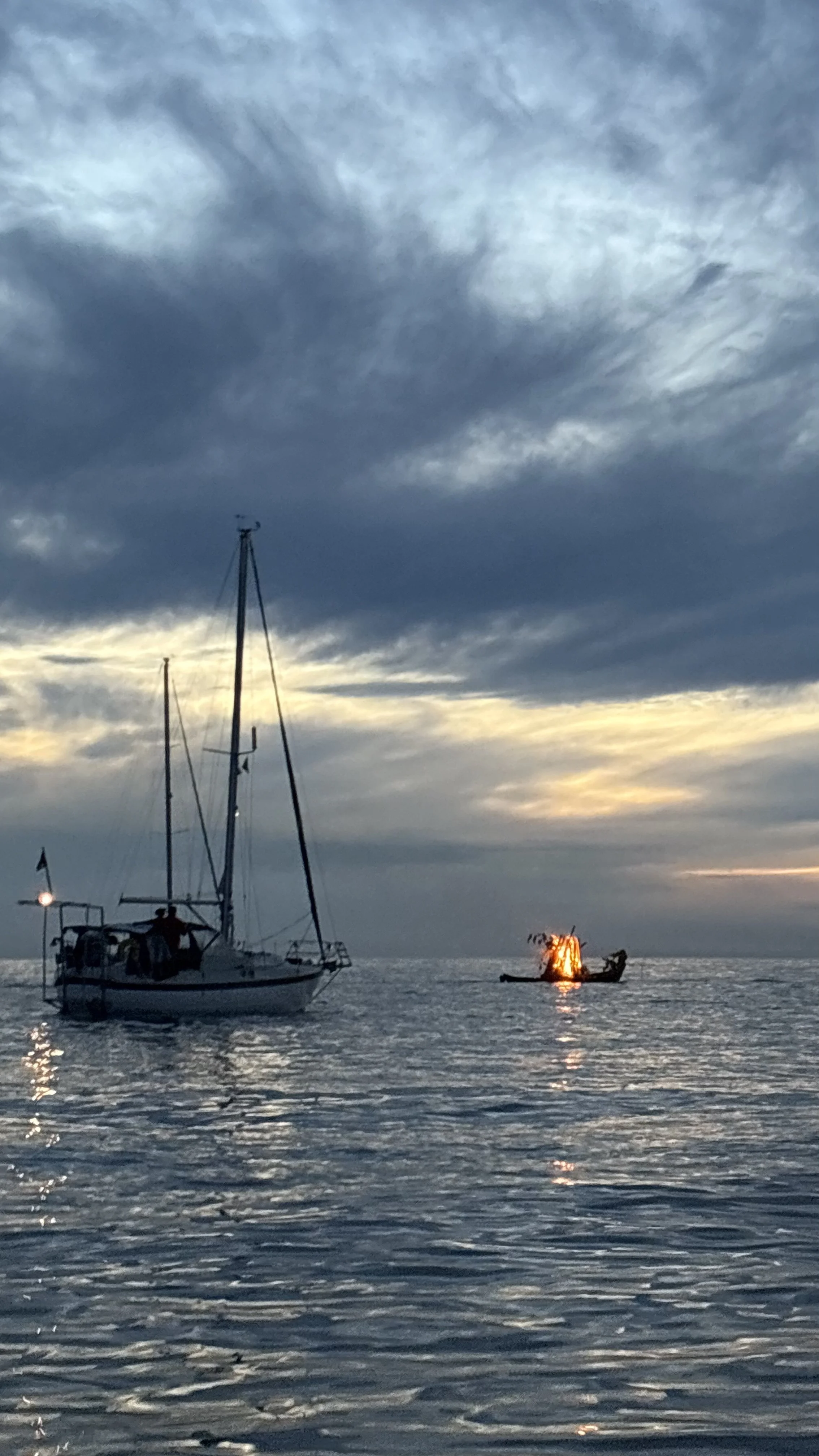A boat sailing on the water during sunset with dark clouds overhead and a small fire on a boat in the distance.