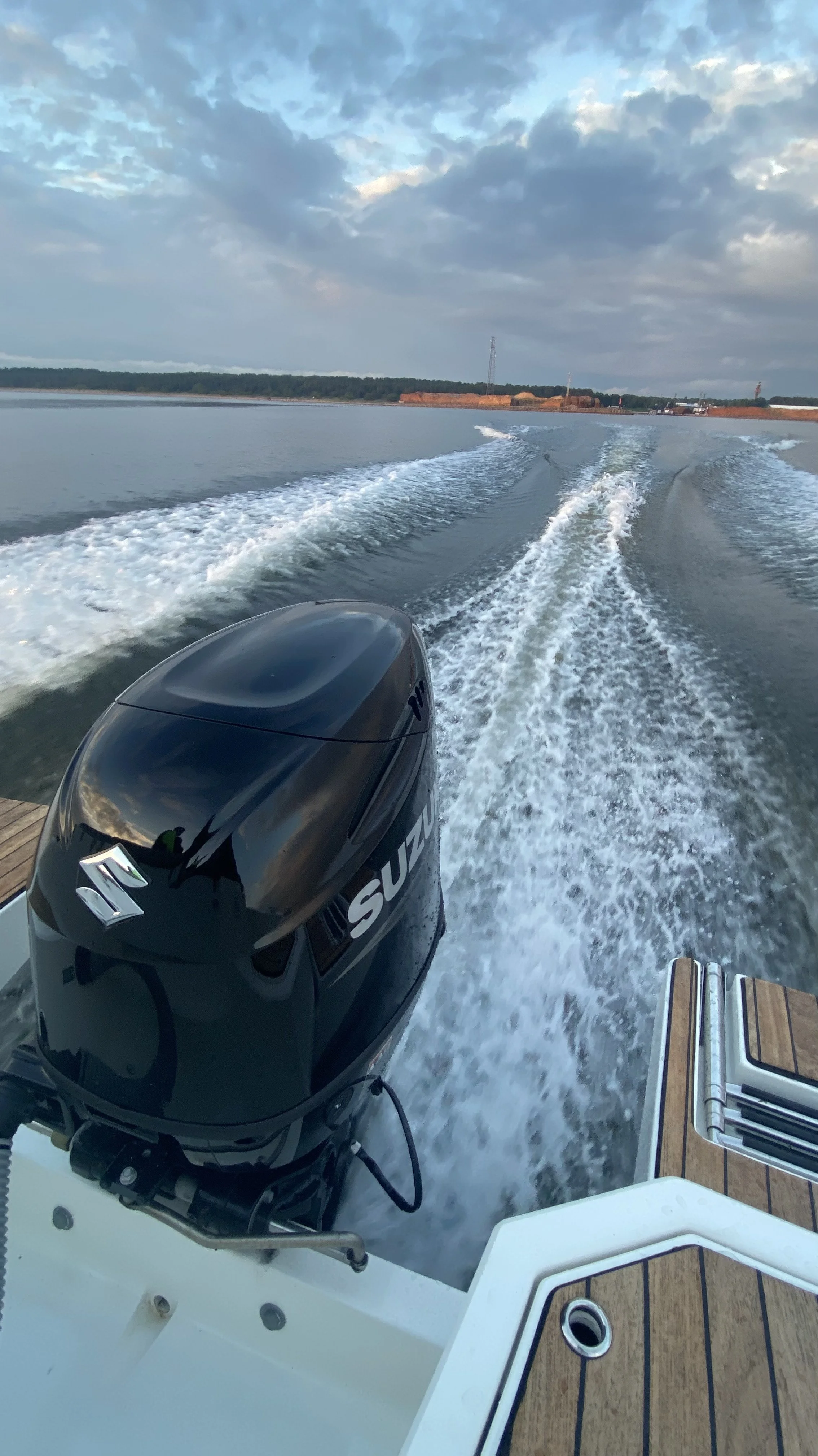 View from the back of a boat showing a Suzuki outboard motor, water trail behind the boat, distant shoreline, and cloudy sky.