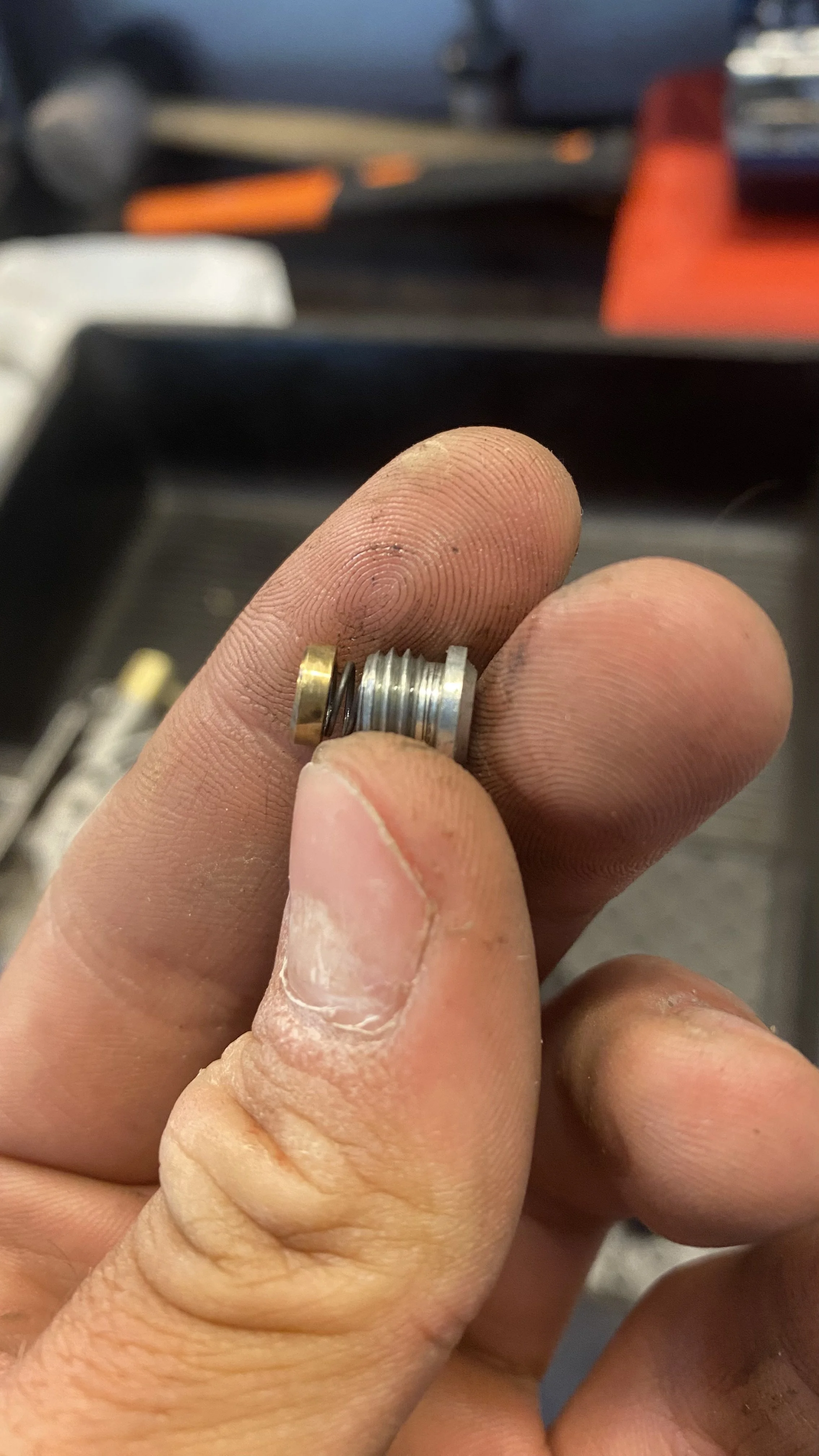Close-up of a person's hand holding a small metal and brass component, with a workbench and tools in the background.