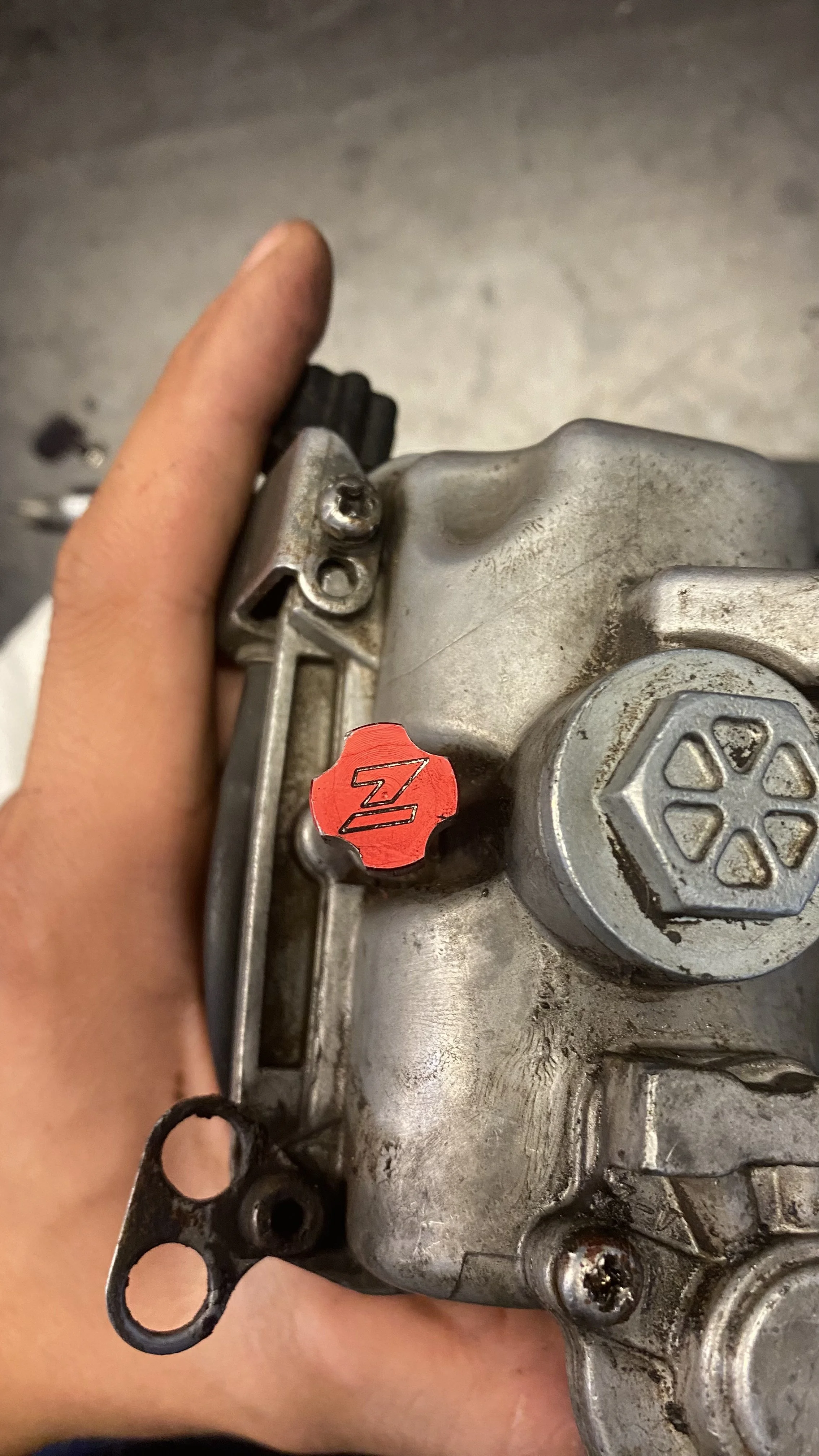 A hand holding a metallic engine component, with a red sticker marked 'Z' on it and visible screws and bolts.