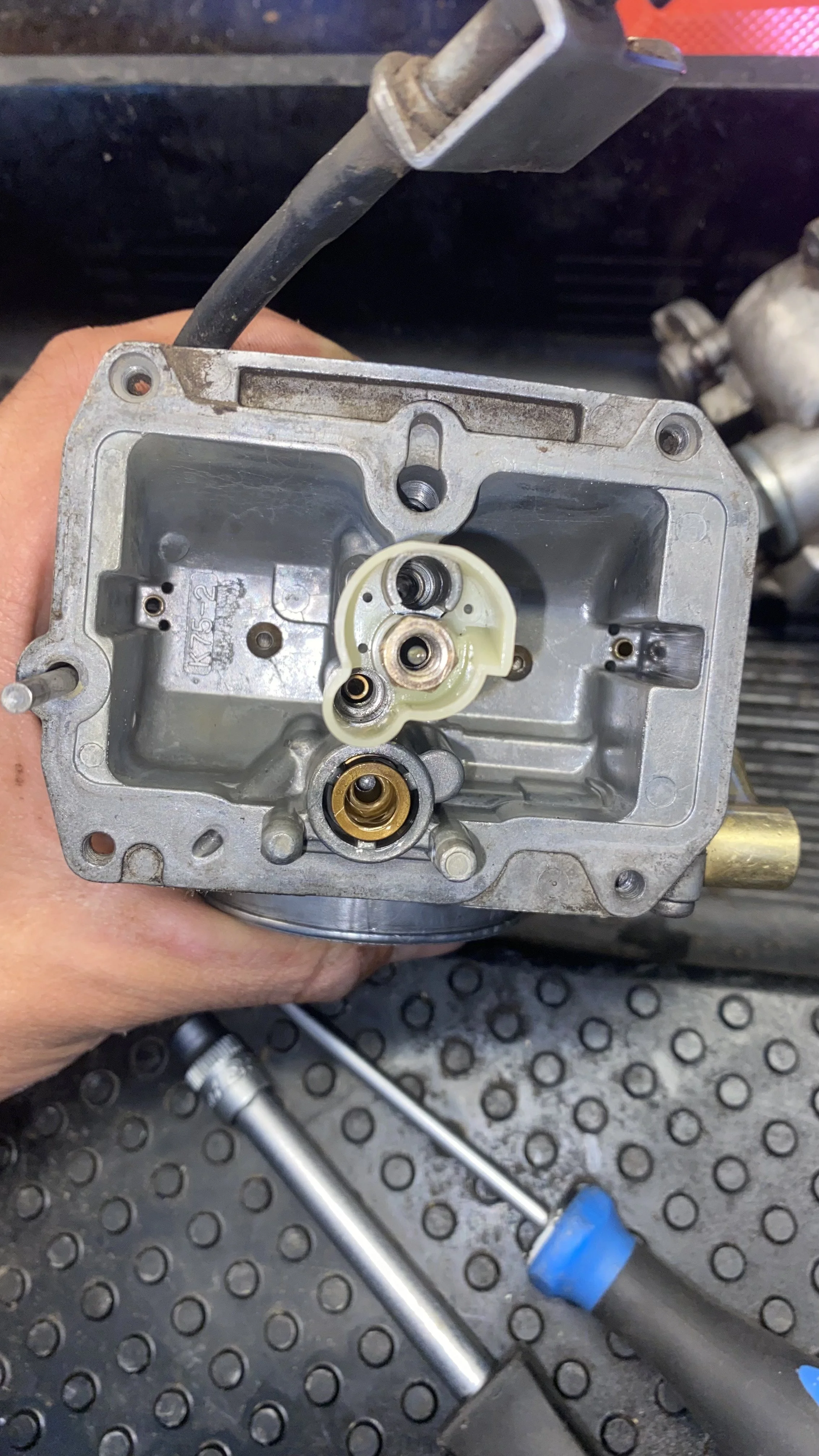 Close-up of a disassembled carburetor, with a person's hand holding it and a screwdriver nearby, on a workbench.