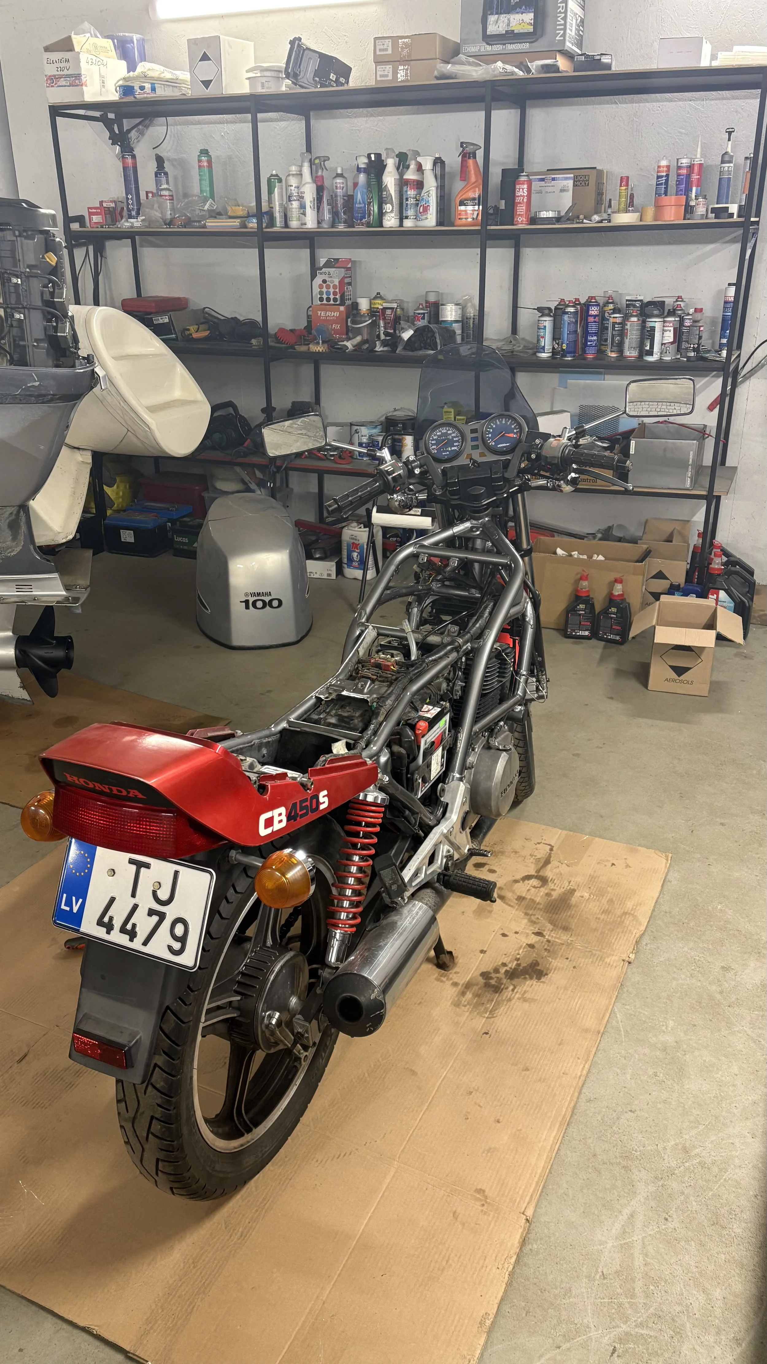 A partially assembled motorcycle in a garage with tools and supplies on shelves in the background. The motorcycle has a red rear section with the label "CB 450S" and a Latvian license plate, and it is placed on a piece of cardboard on a concrete floo