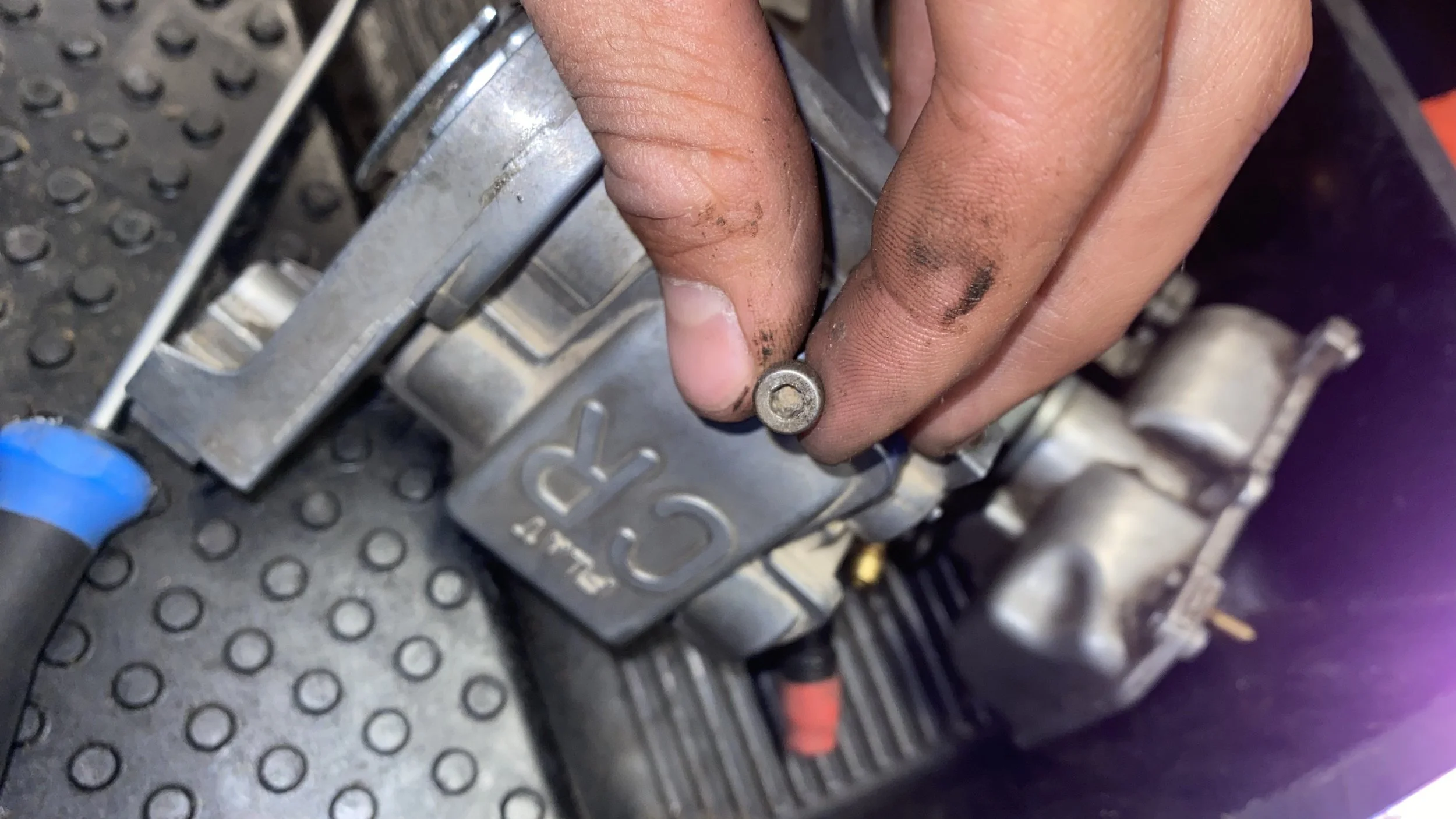 Close-up of a person's hand with dirty fingers holding a small screw or bolt near a metallic component, likely part of a mechanical or electronic device.