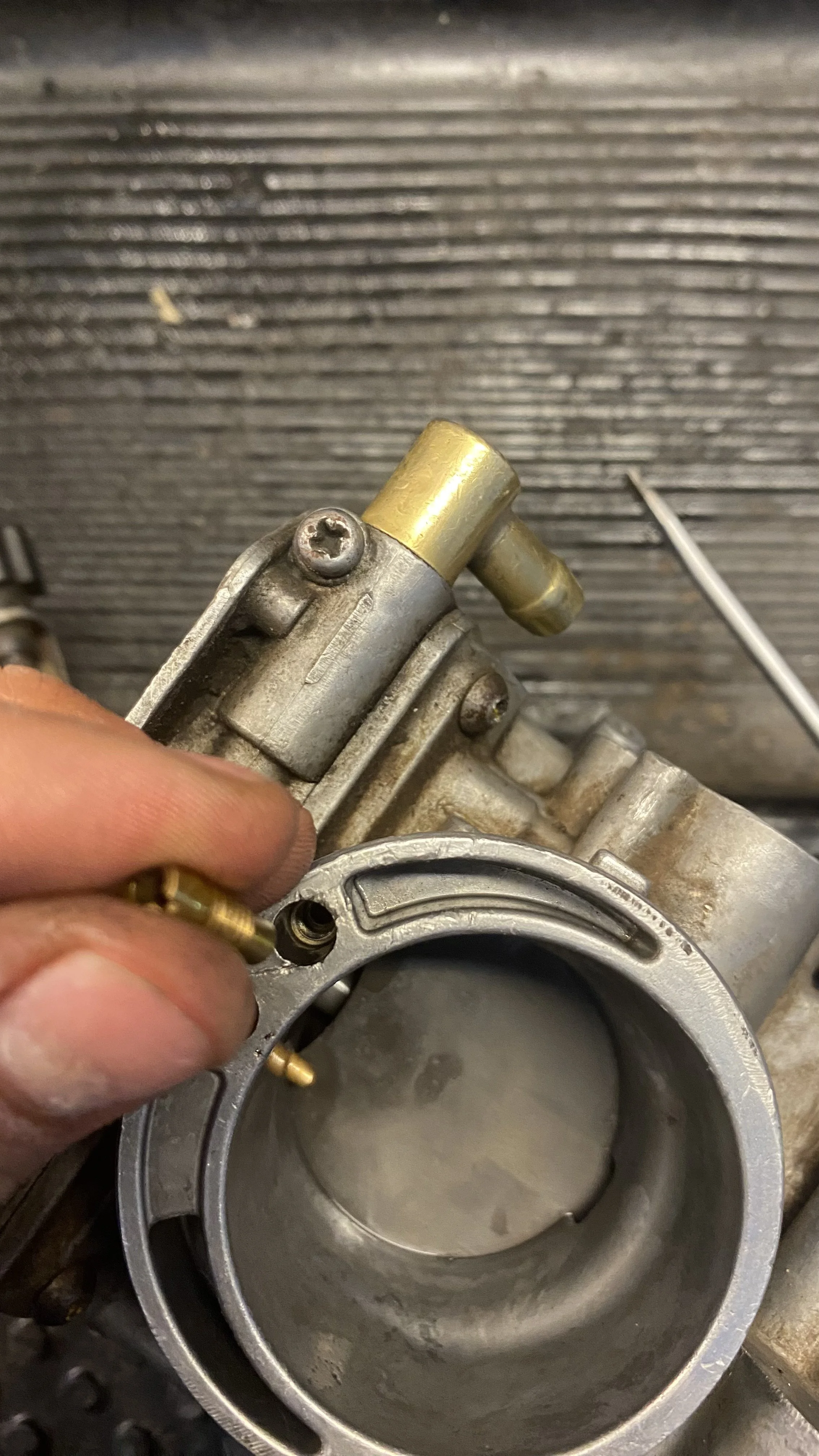 Close-up of a carburetor with a person's fingers holding a small screw. The carburetor has metallic parts, some brass-colored components, and a large circular opening at the bottom. The background is a textured, dark surface.