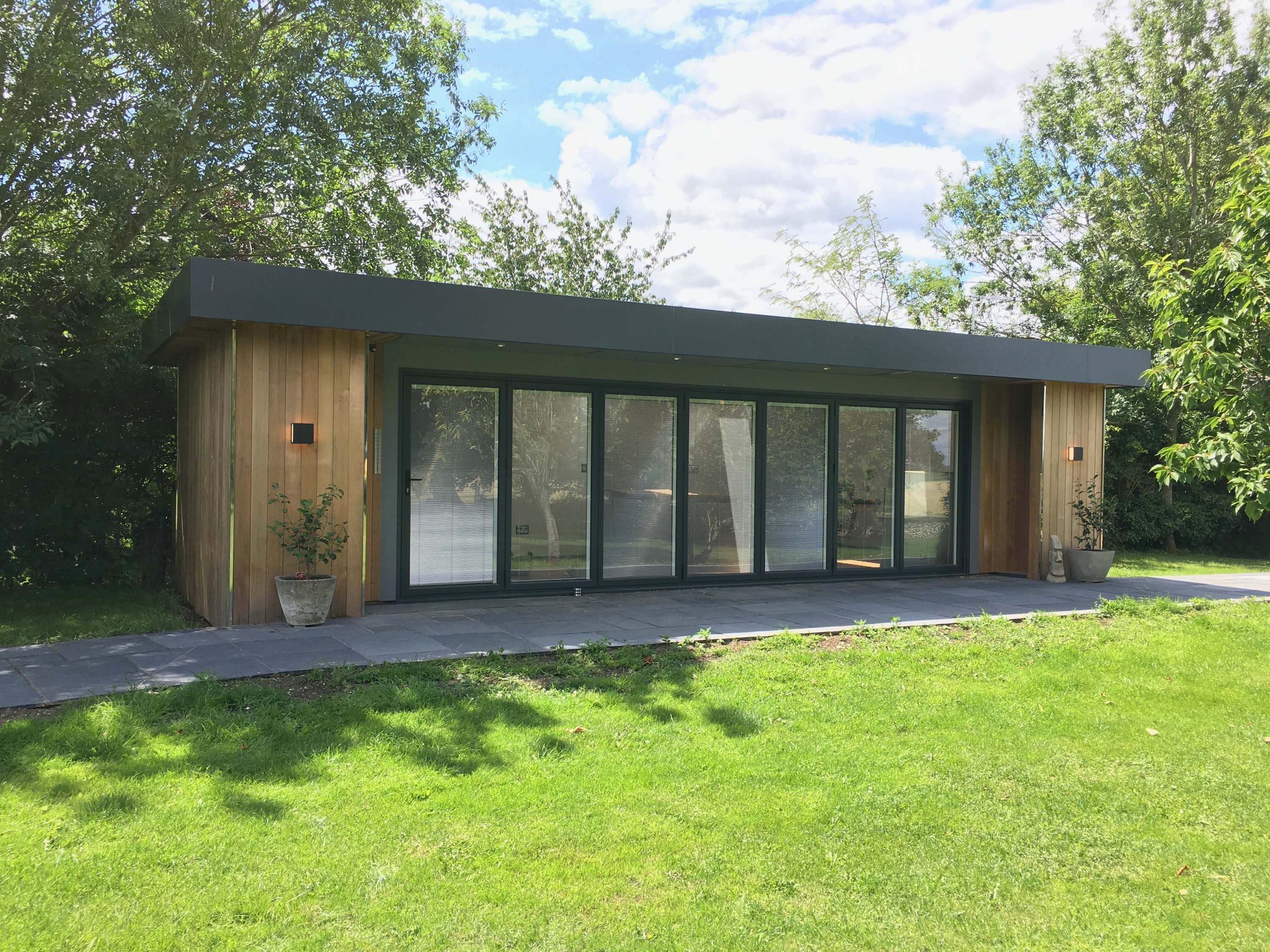 Modern house with large glass doors, wooden exterior, and a flat roof, surrounded by green trees and lawn.