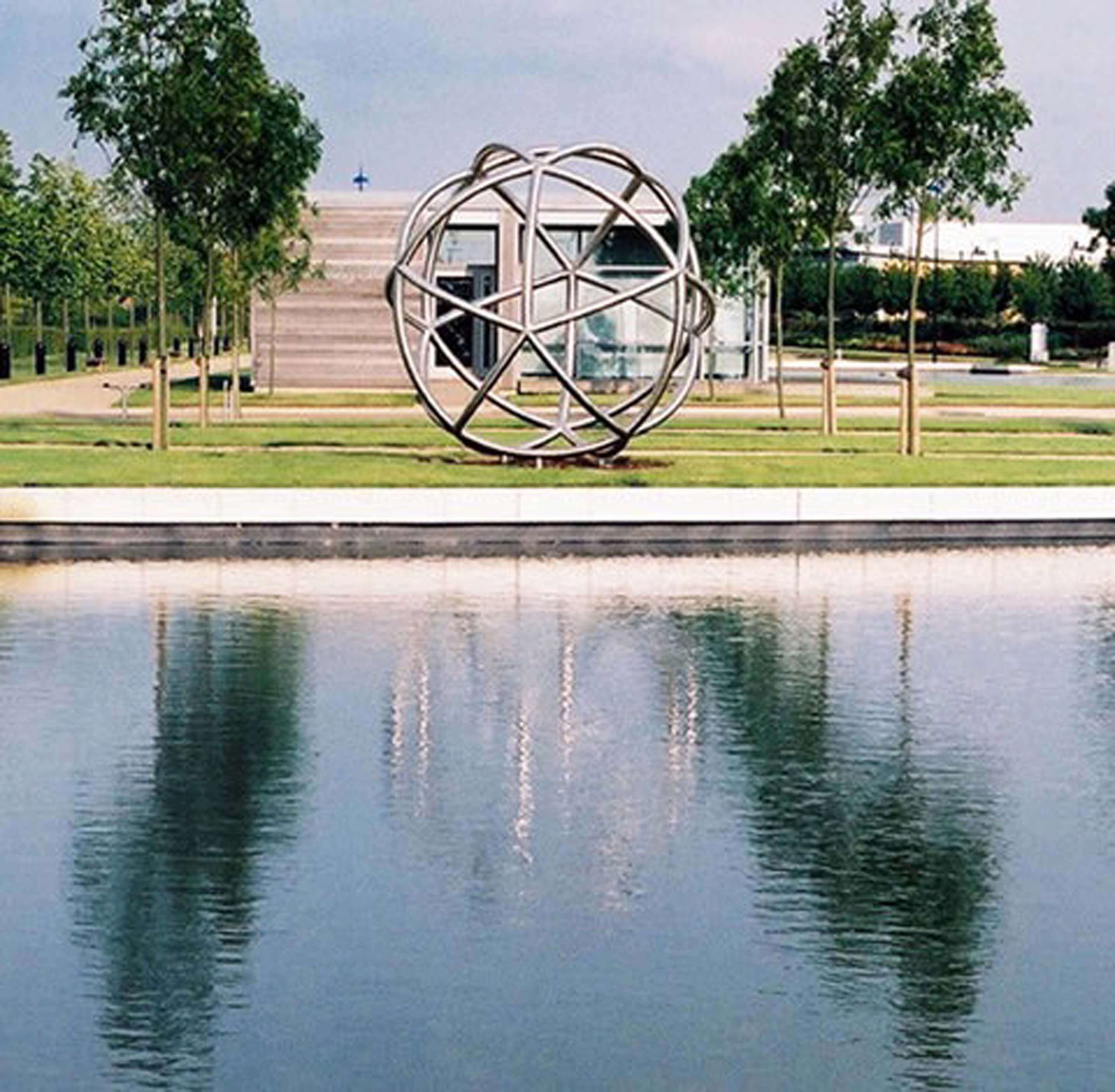 A park scene featuring a decorative spherical metal sculpture near a body of water, with trees, a modern building, and a cloudy sky in the background.