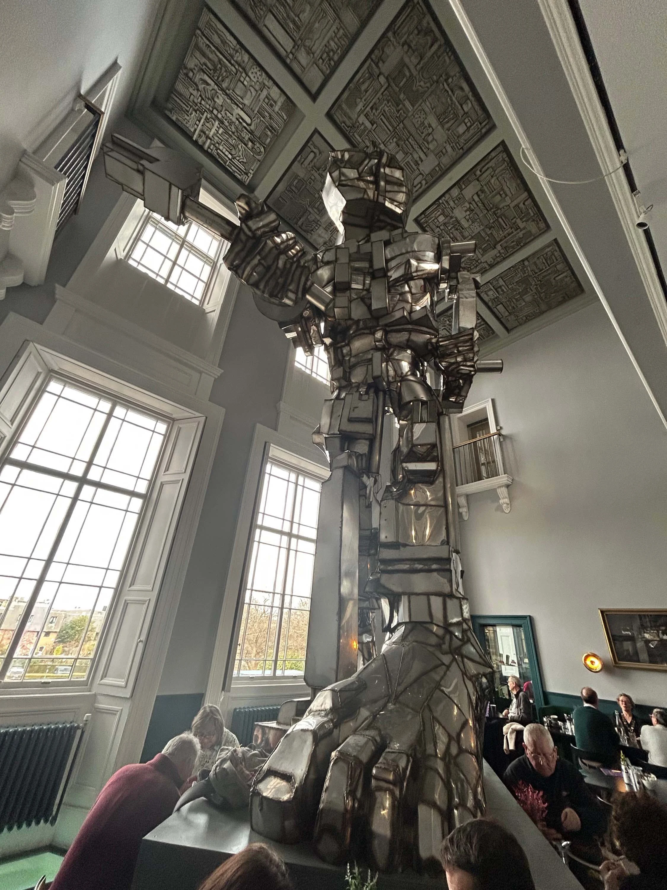People sitting in a room with large windows, a radiator, and artwork on the walls, while a large metallic sculpture of a human figure is in the center.