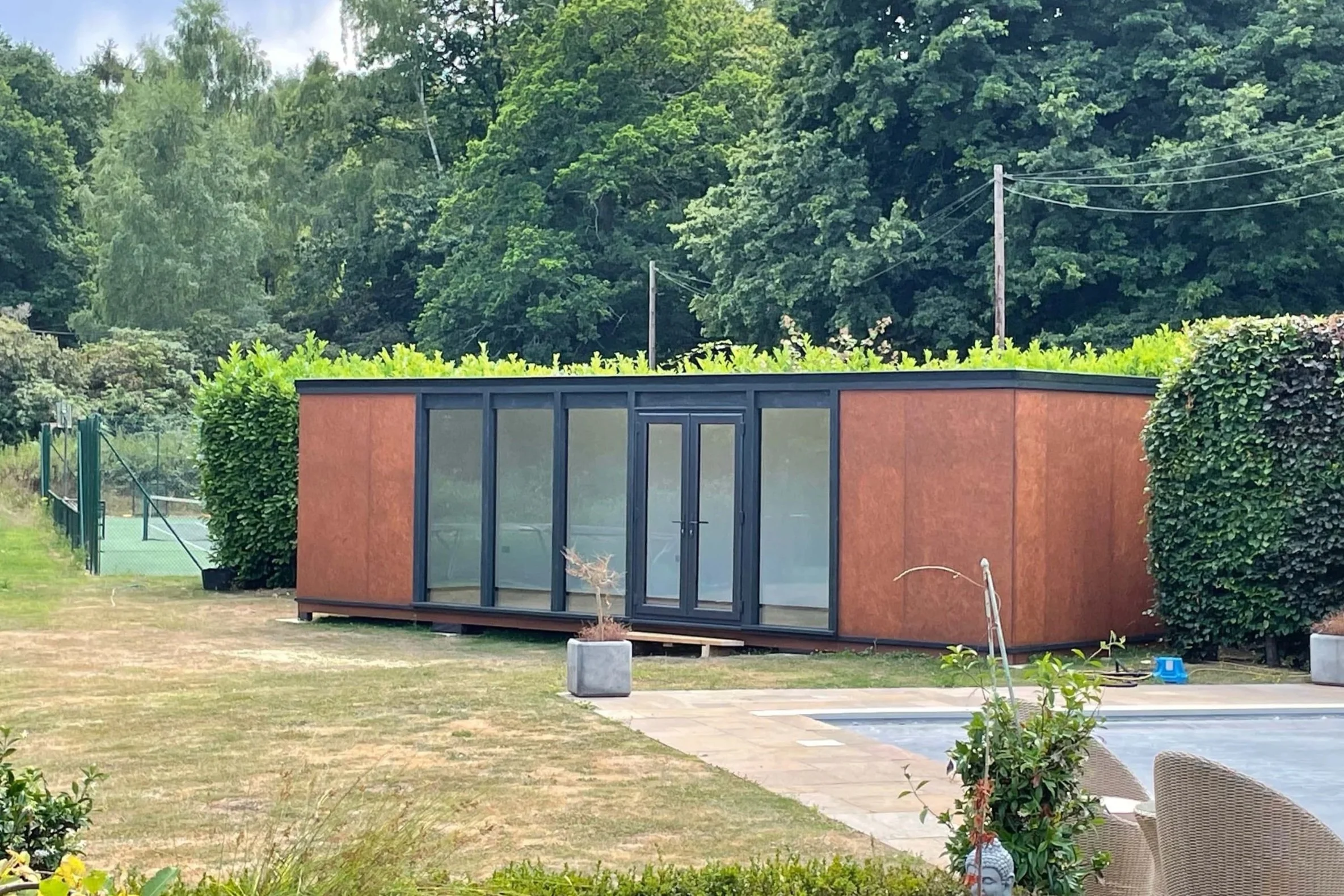 A modern, minimalist house with large glass doors and a flat roof, surrounded by lush green trees and a well-maintained yard with some small plants and outdoor furniture, under a partly cloudy sky.
