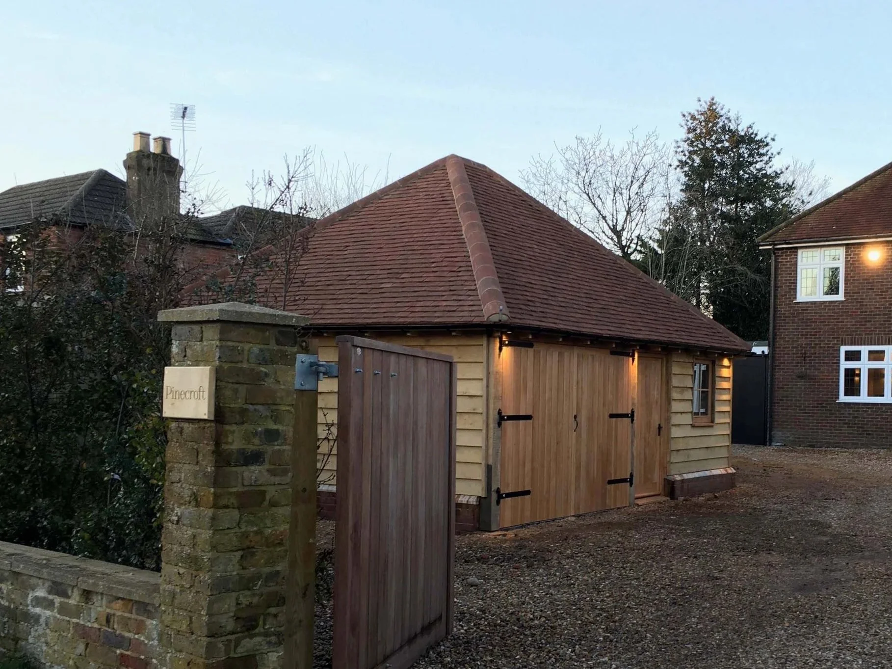 A small wooden shed with black hinges and a sloped red tile roof, located in a backyard near brick houses. A brick pillar with a sign reading 'Pinecroft' is in the foreground, and there are trees and houses in the background.