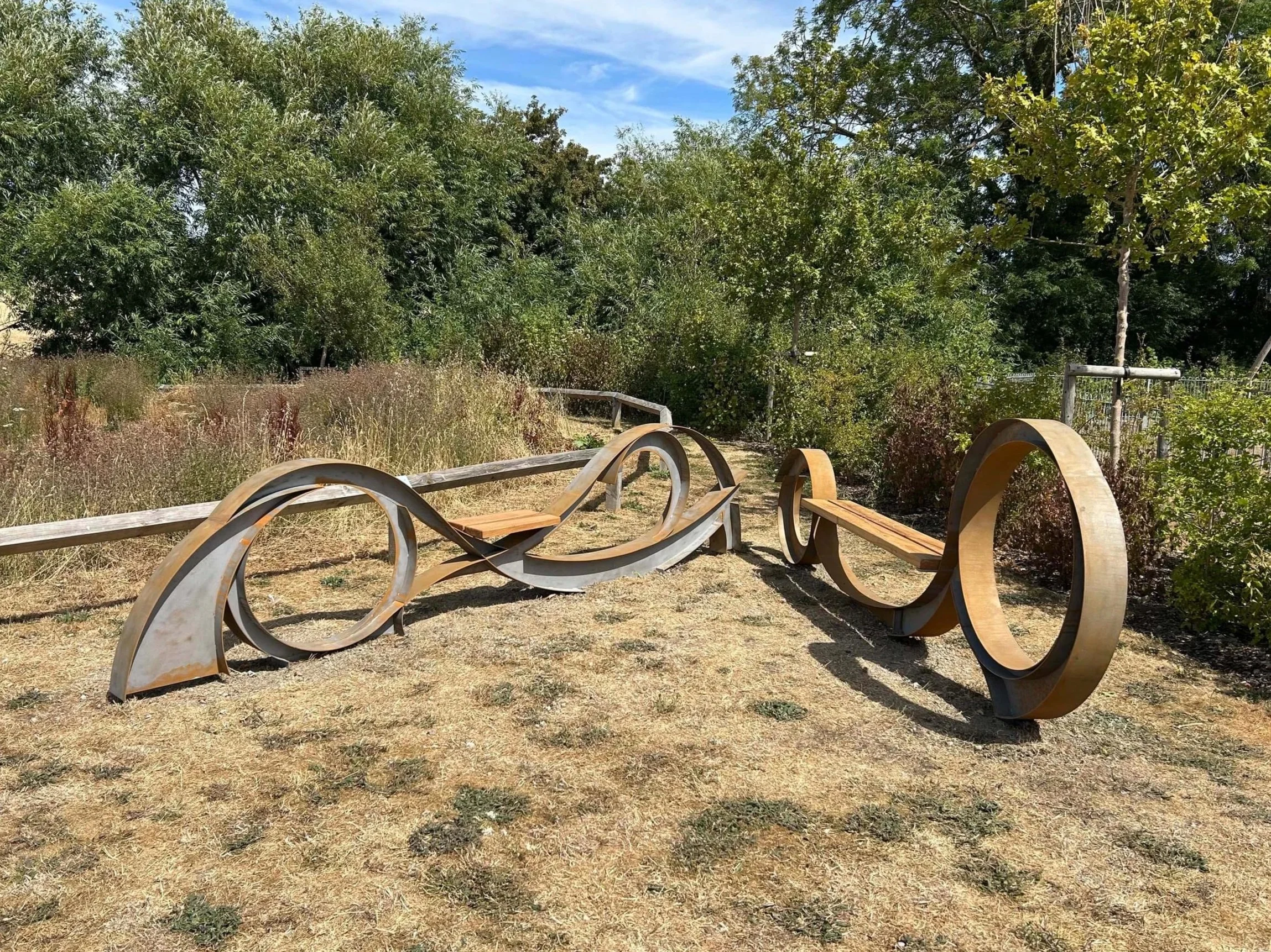A park with a wooden sculpture made of curved, intertwined wooden planks on dry grass, surrounded by green trees and bushes, under a blue sky with some clouds.