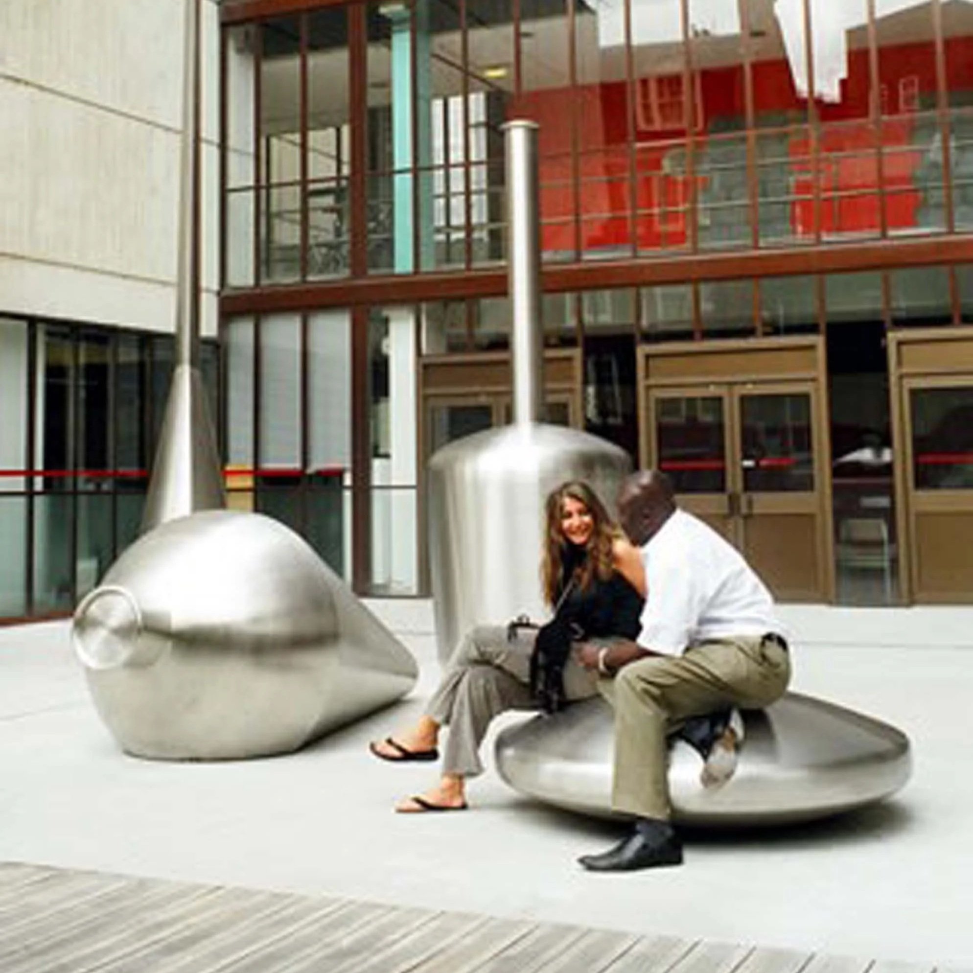Two women sitting on the floor, smiling, with their heads close together, in a modern building with large glass windows and red and beige exterior design.