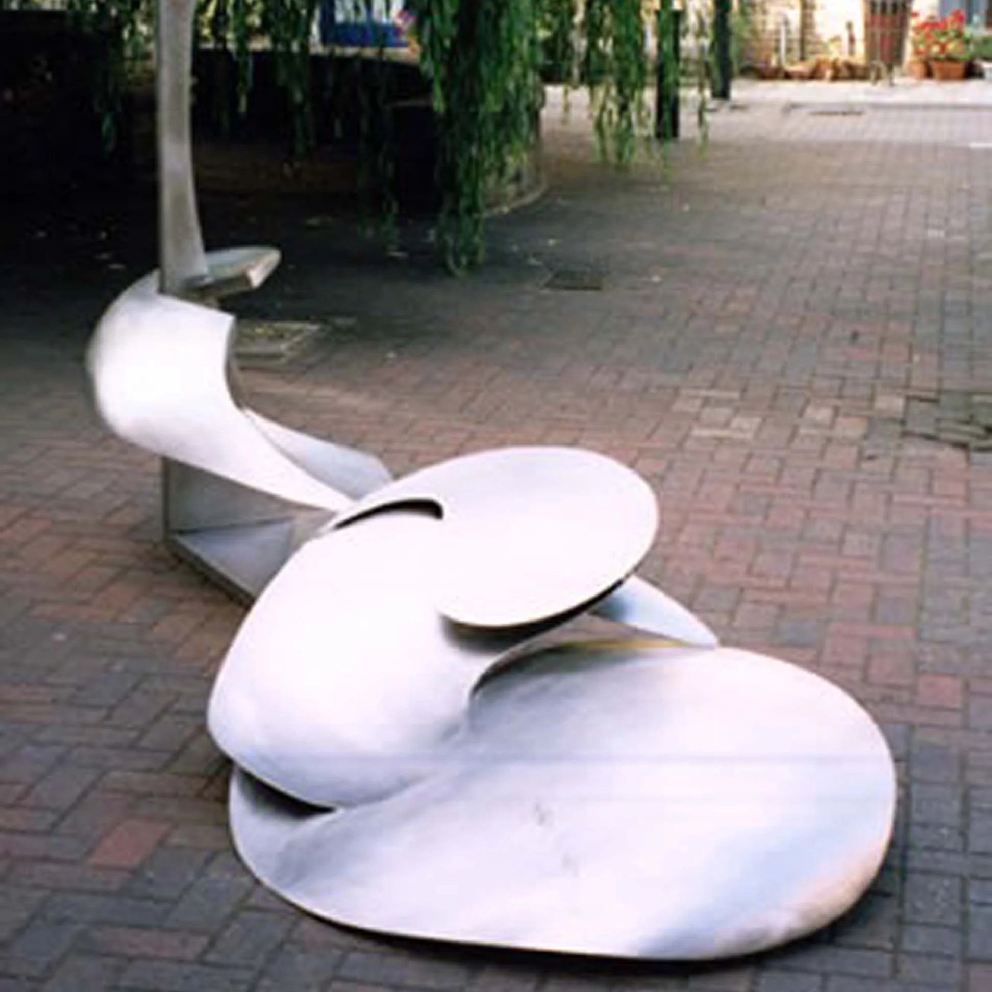 Outdoor scene with white modern lounge chairs arranged on a brick-paved surface, some greenery in the background, possibly in a park or plaza.