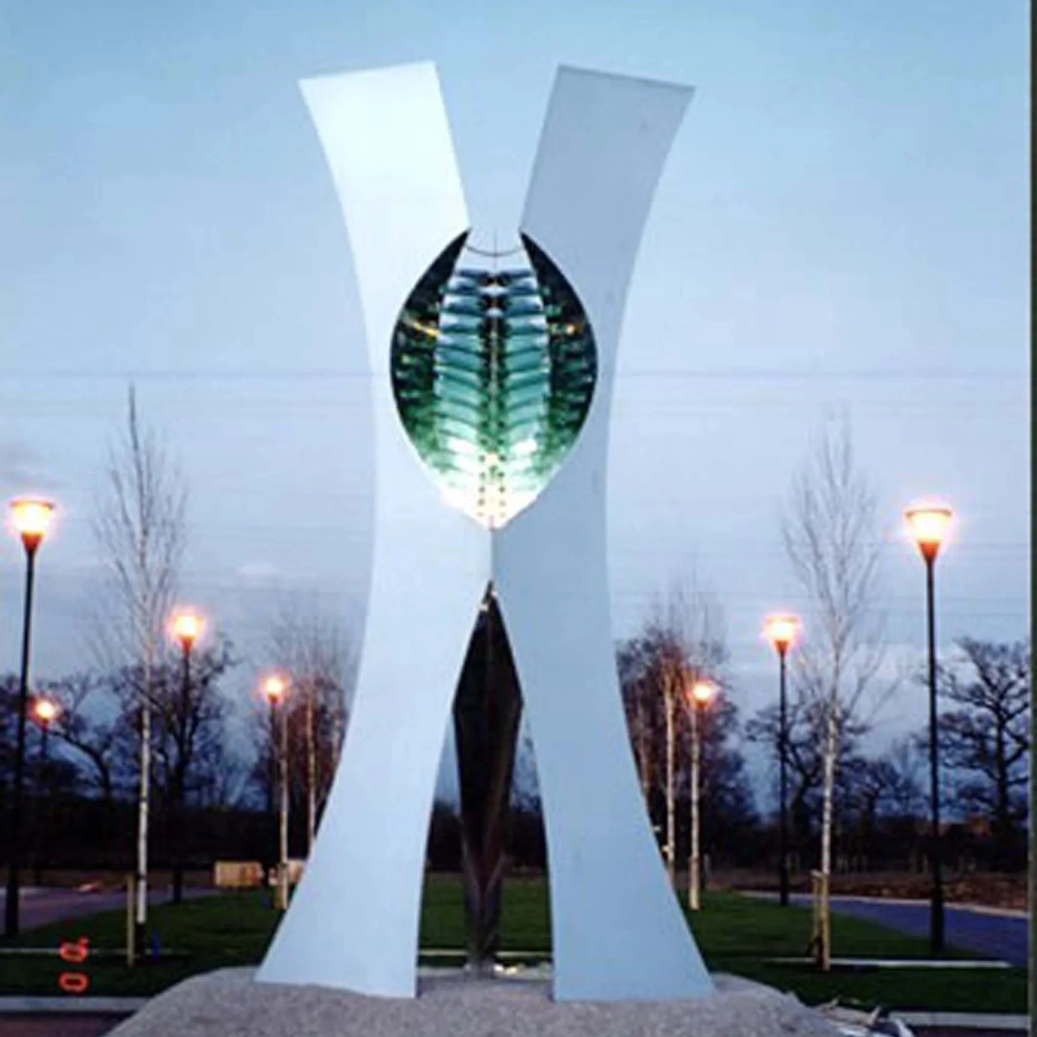 A large, modern sculpture of a stylized human face with black and silver metallic elements, set outdoors against a cloudy sky with leafless trees in the background.