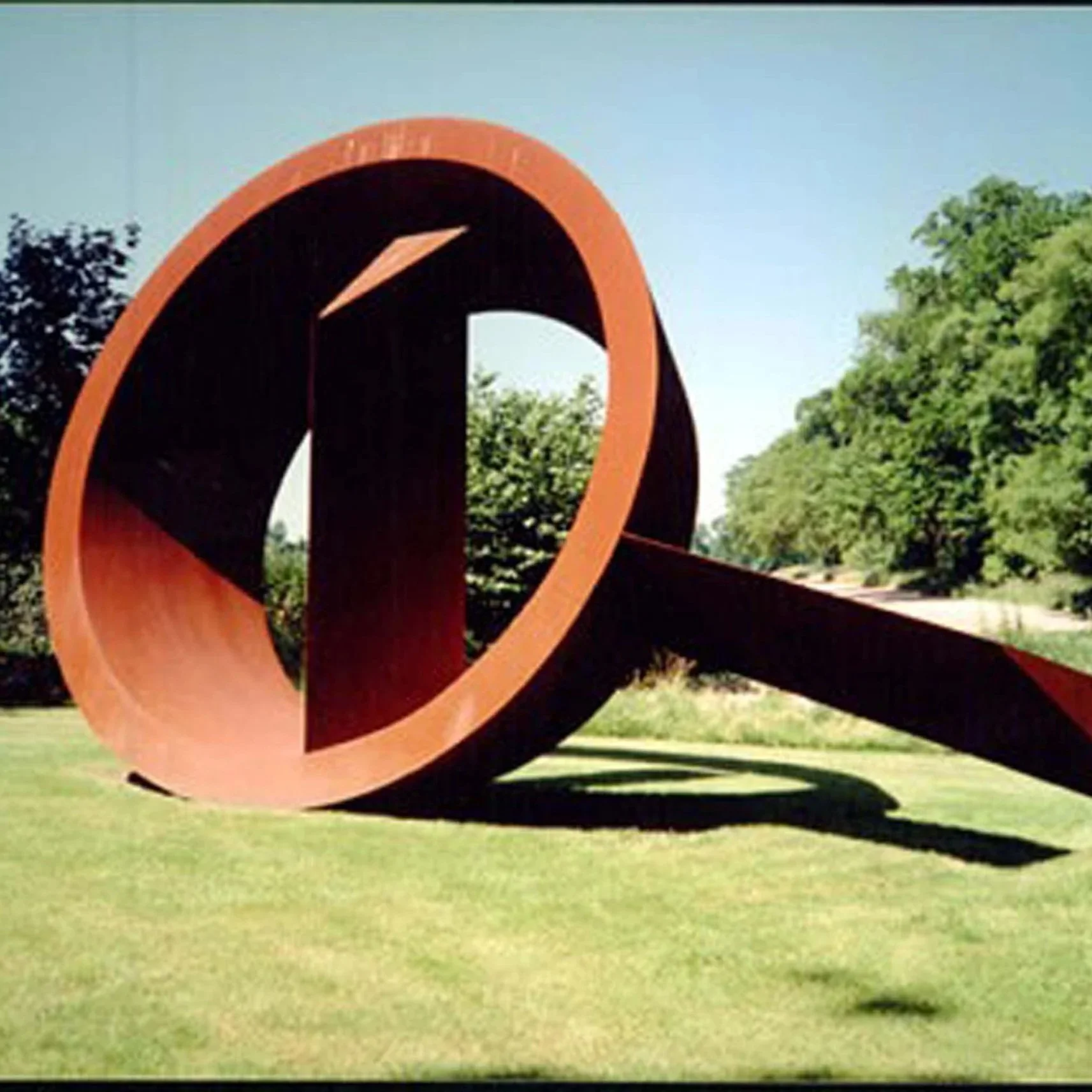 Large abstract outdoor sculpture with a rolled shape, painted in orange and black, situated on a grassy area with trees and clear blue sky in the background.