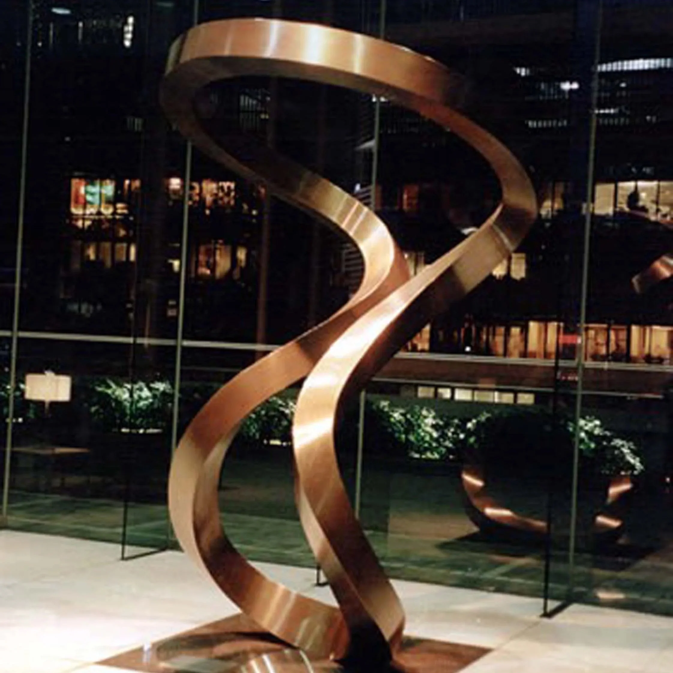 A large metal sculpture of a spiral made of shiny, reflective metal in an indoor setting with a dark background and some greenery at the bottom.