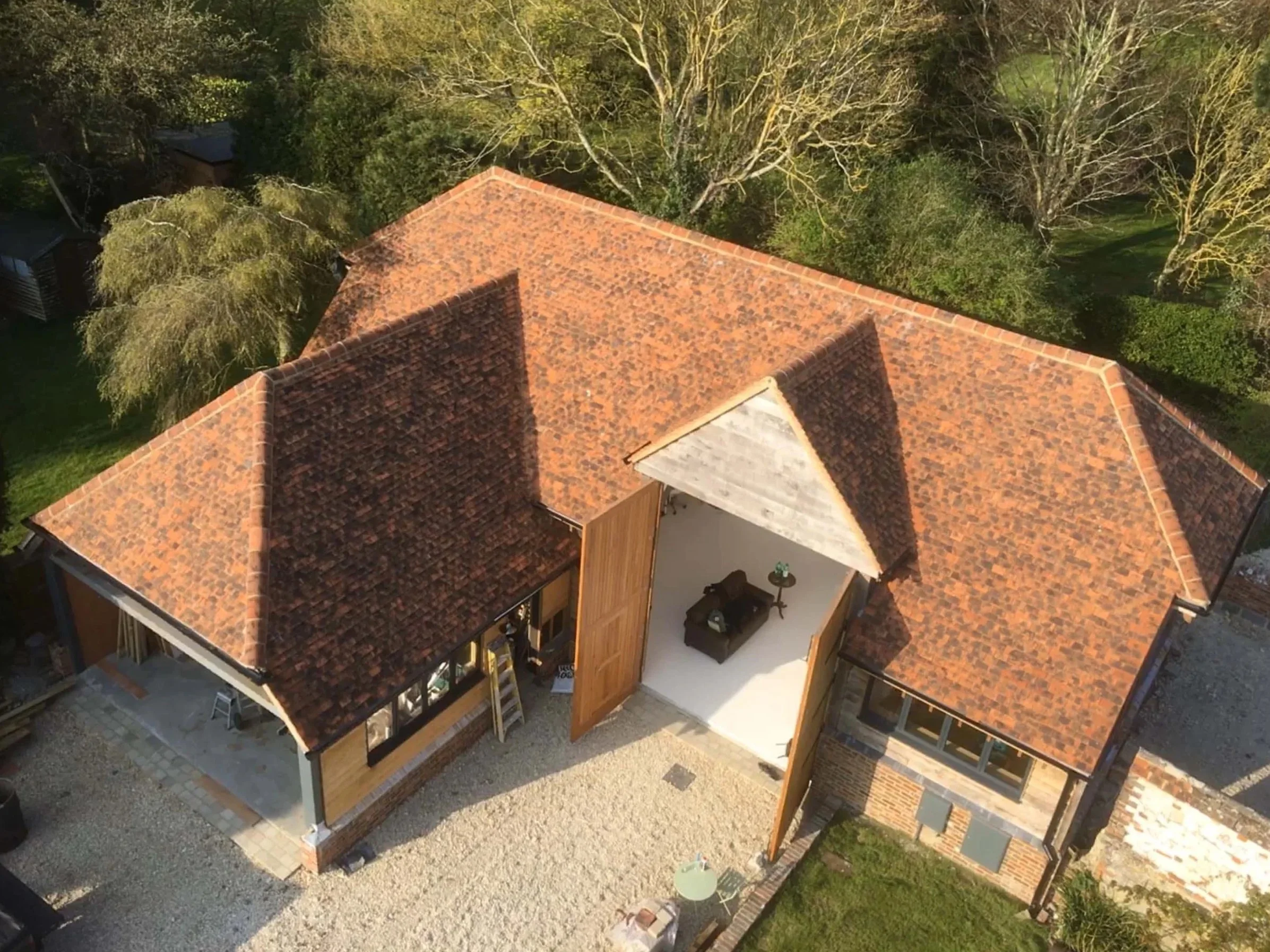 Aerial view of a house under construction with a brown tiled roof, surrounded by trees and a gravel yard.