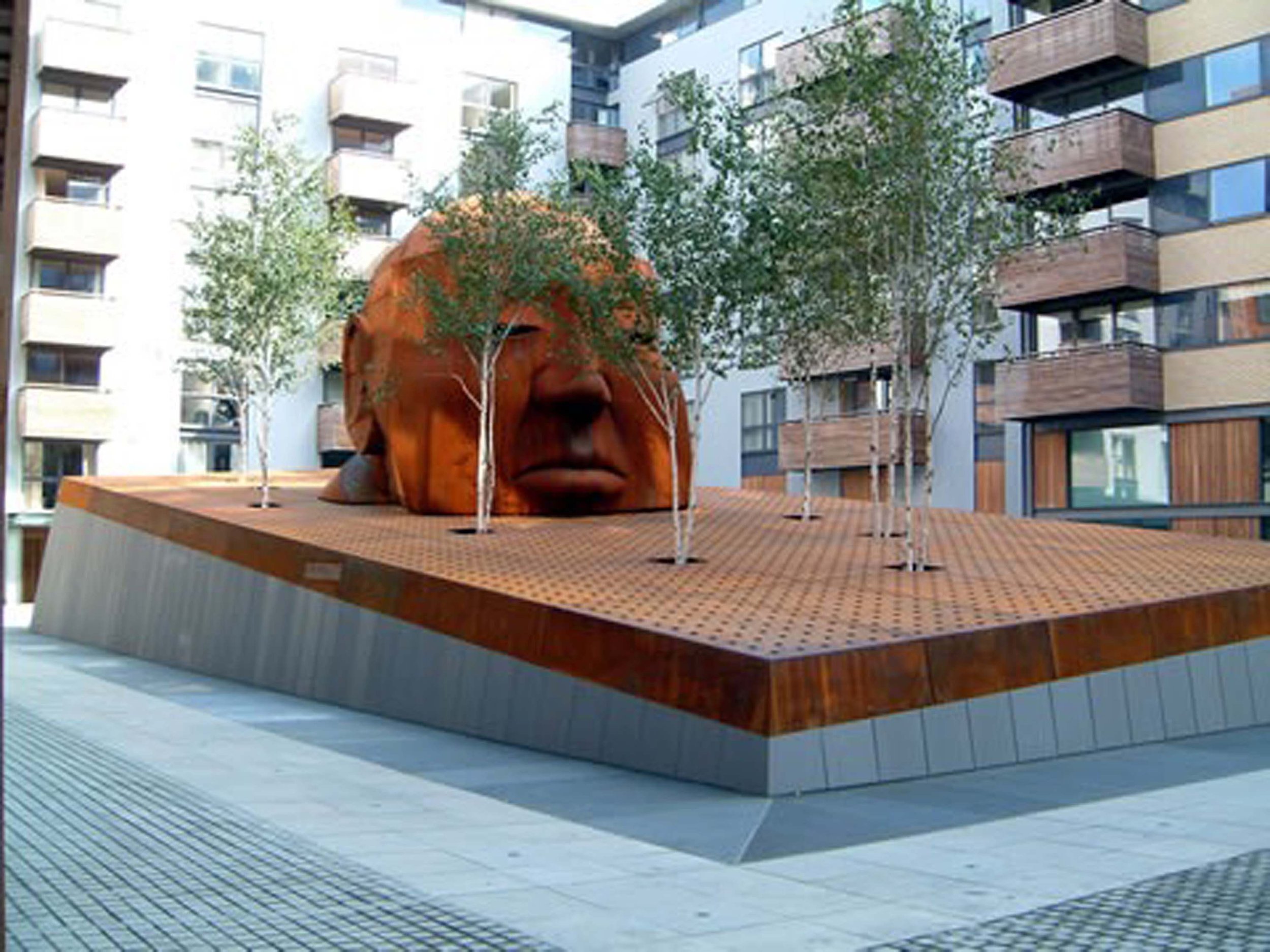 Outdoor courtyard with a large artistic sculpture resembling a human face with trees growing around it, surrounded by modern apartment buildings.