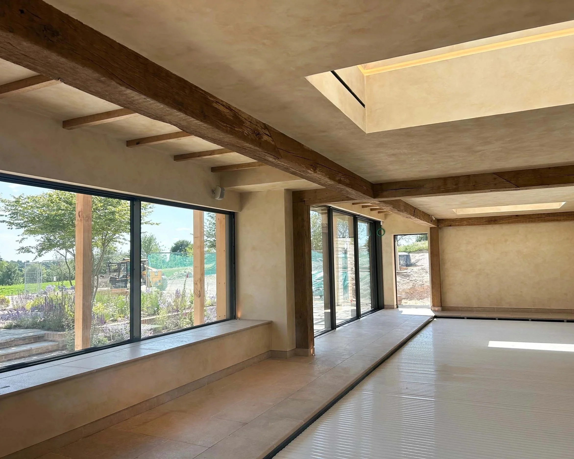 Interior of a modern room with large glass windows, wooden beams on the ceiling, and a light-colored tiled floor, with a view of trees outside.