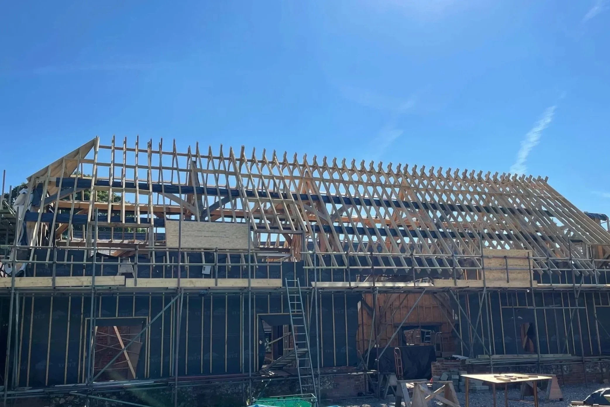 Construction site with wooden framing for the roof of a building, scaffolding surrounding the structure, and a bright blue sky overhead.