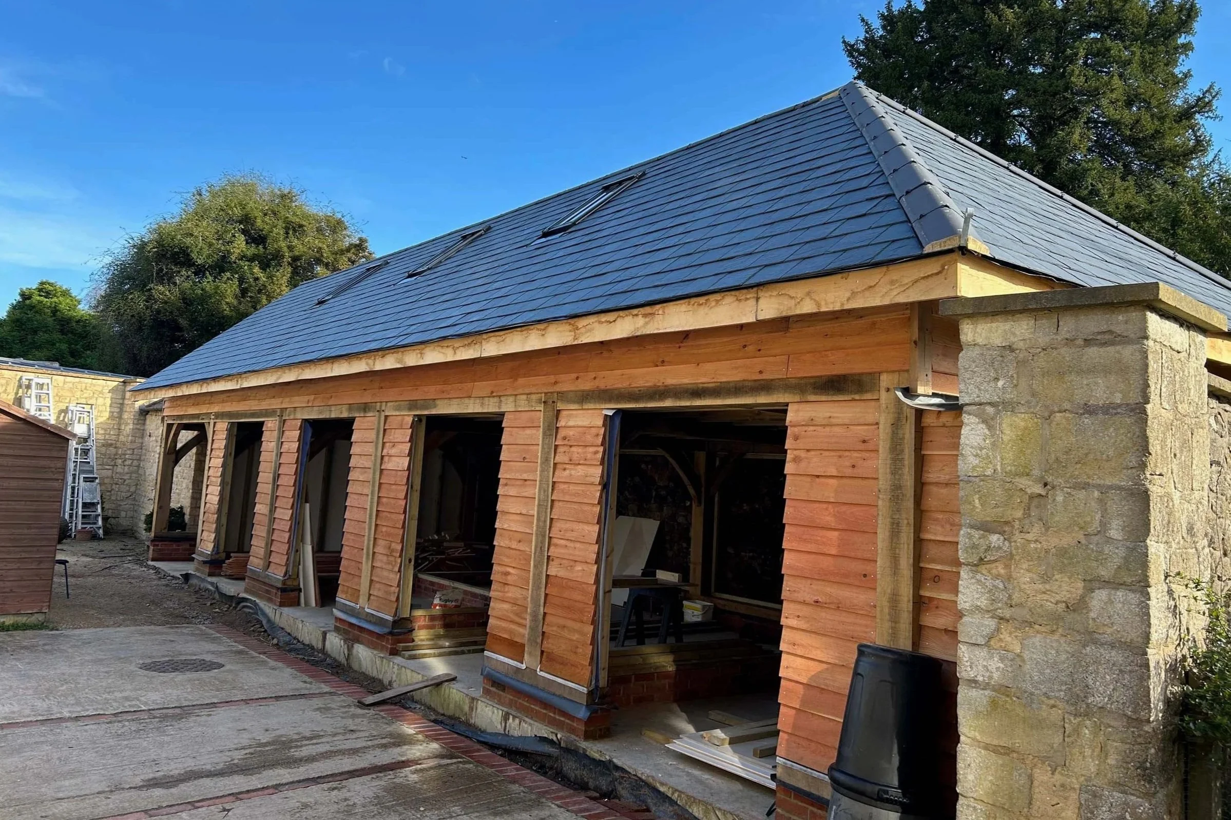Under construction building with a new slate roof and wooden framing, a stone wall, and construction tools and materials nearby, under a blue sky with trees in the background.