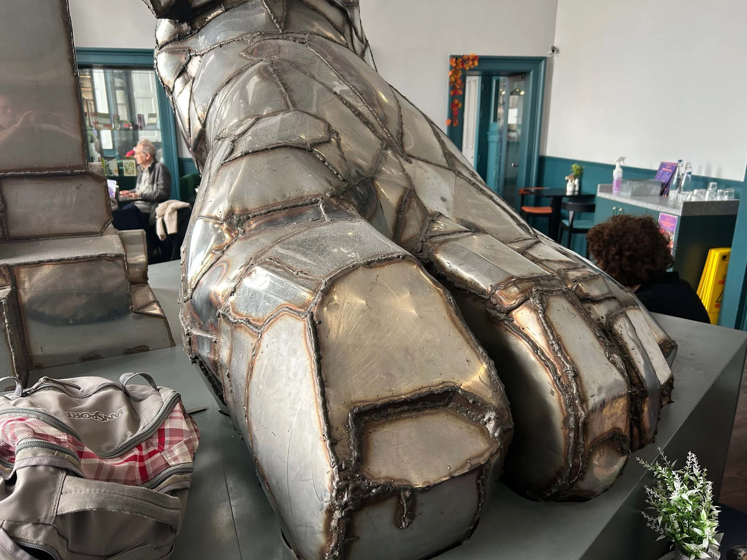 Silver metallic sculpture with intricate weld patterns in an indoor space, with a person with curly hair sitting nearby and a woman in the background working on a laptop.