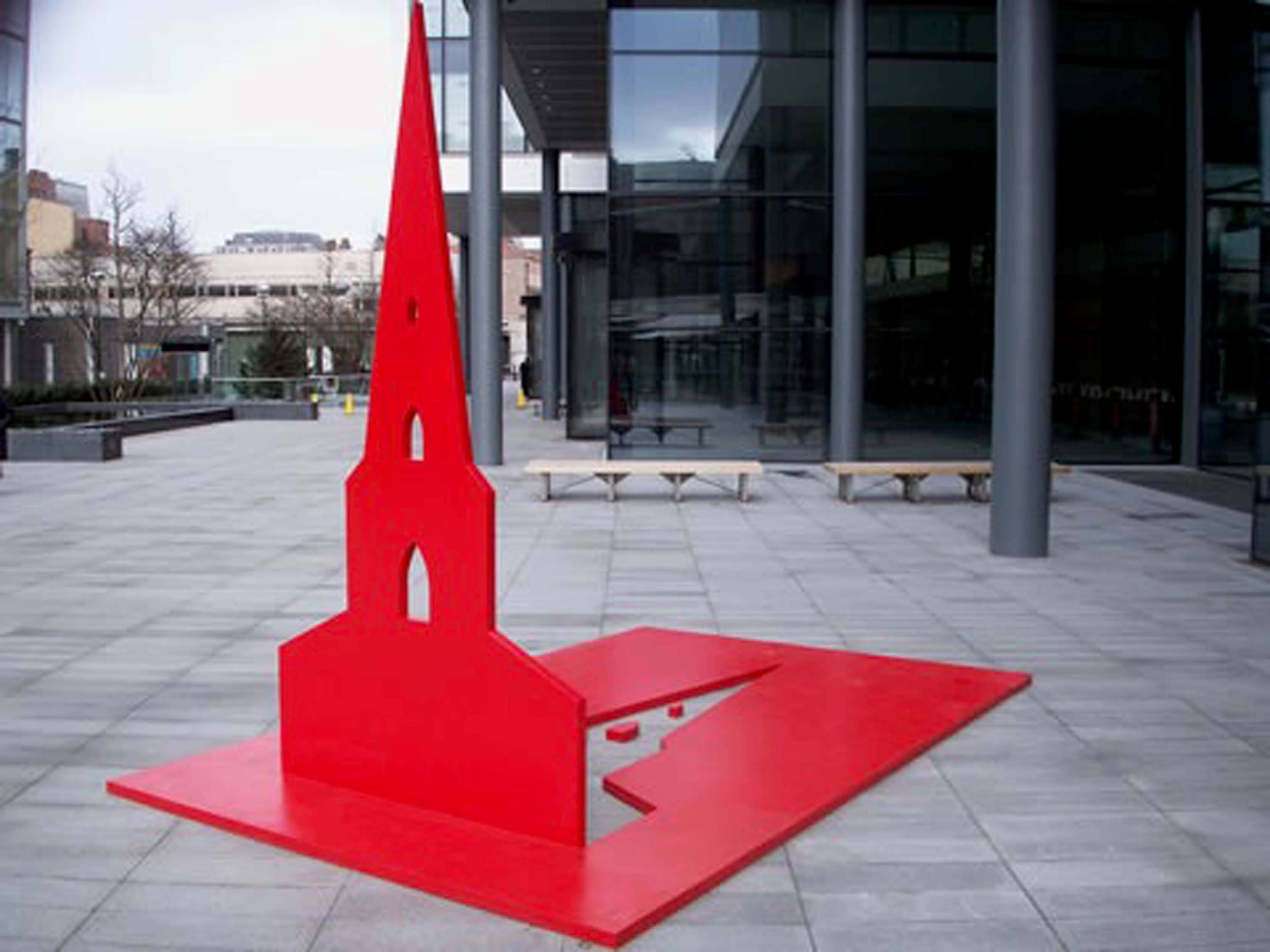 Red chair shaped like a rocket with a tall red fin, placed outdoors near a building with large glass windows, on a paved area with benches and trees in the background.