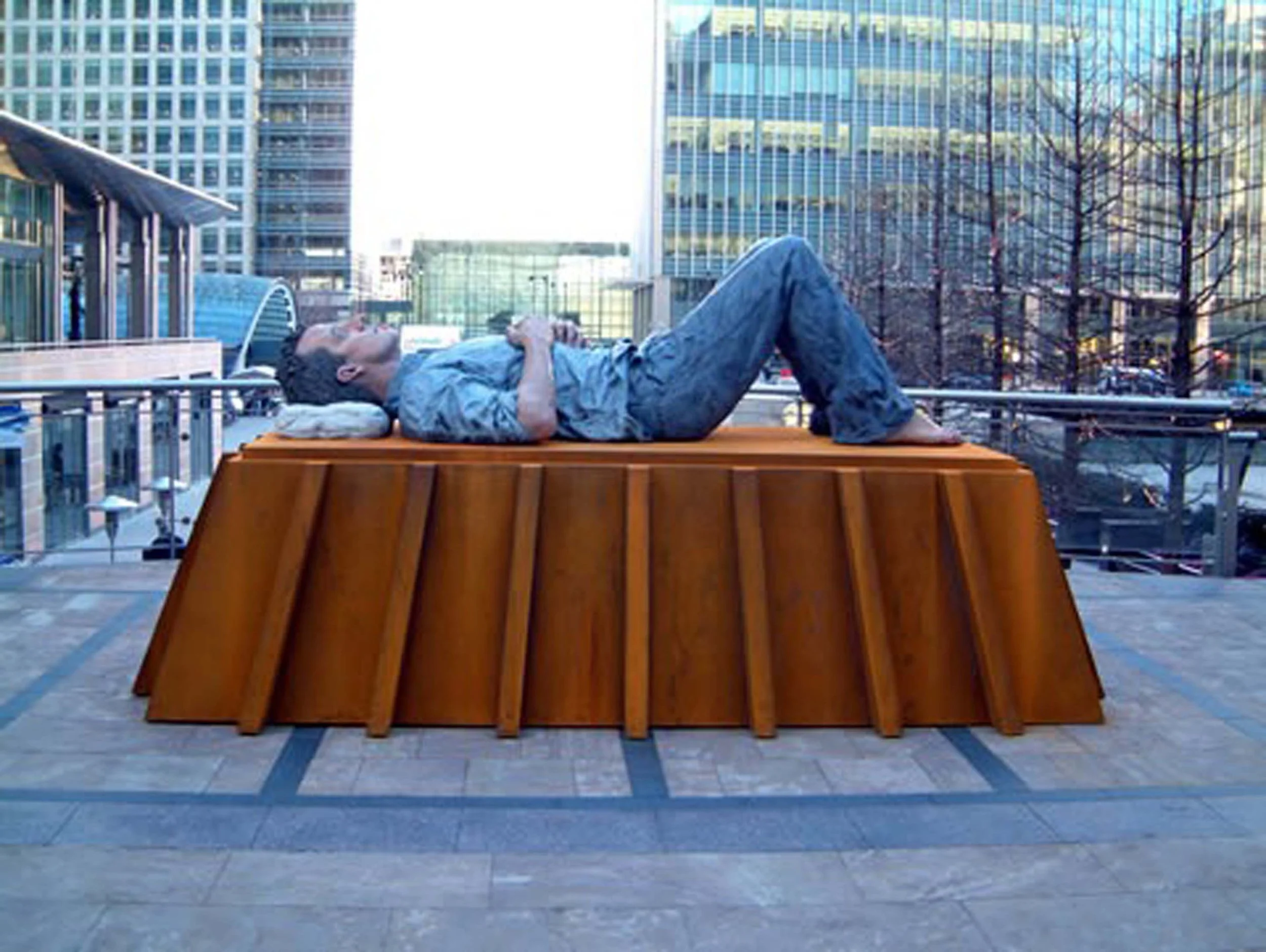 A man lies horizontal on a wooden platform in an urban setting with tall glass buildings and a city street in the background, resting with his eyes closed.