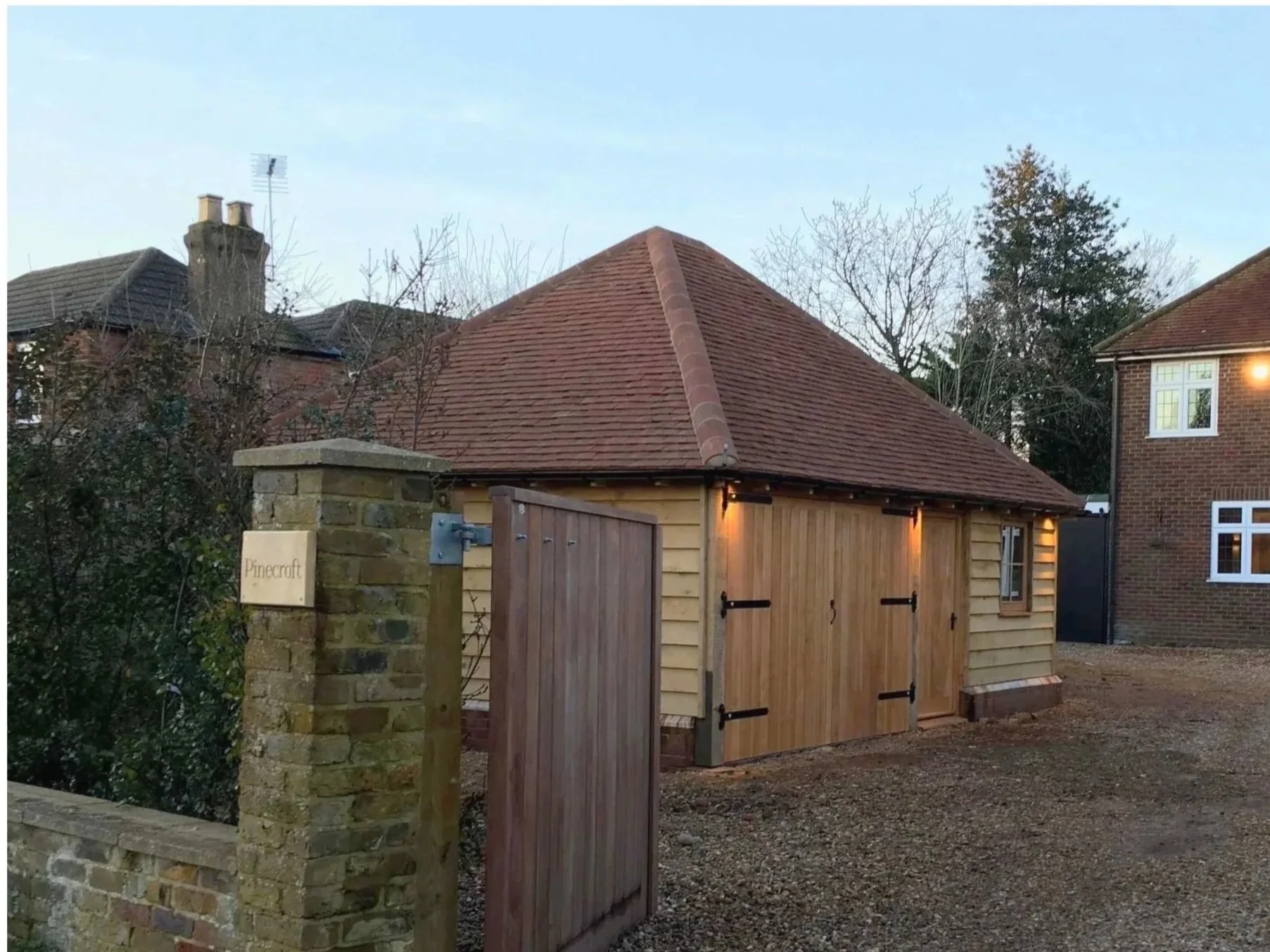 Small wooden shed with a sloped red tile roof, black hinges, and a warm exterior light, situated behind a brick and stone entrance gate with a sign reading 'Pinecroft' during dusk with neighboring houses and trees in the background.
