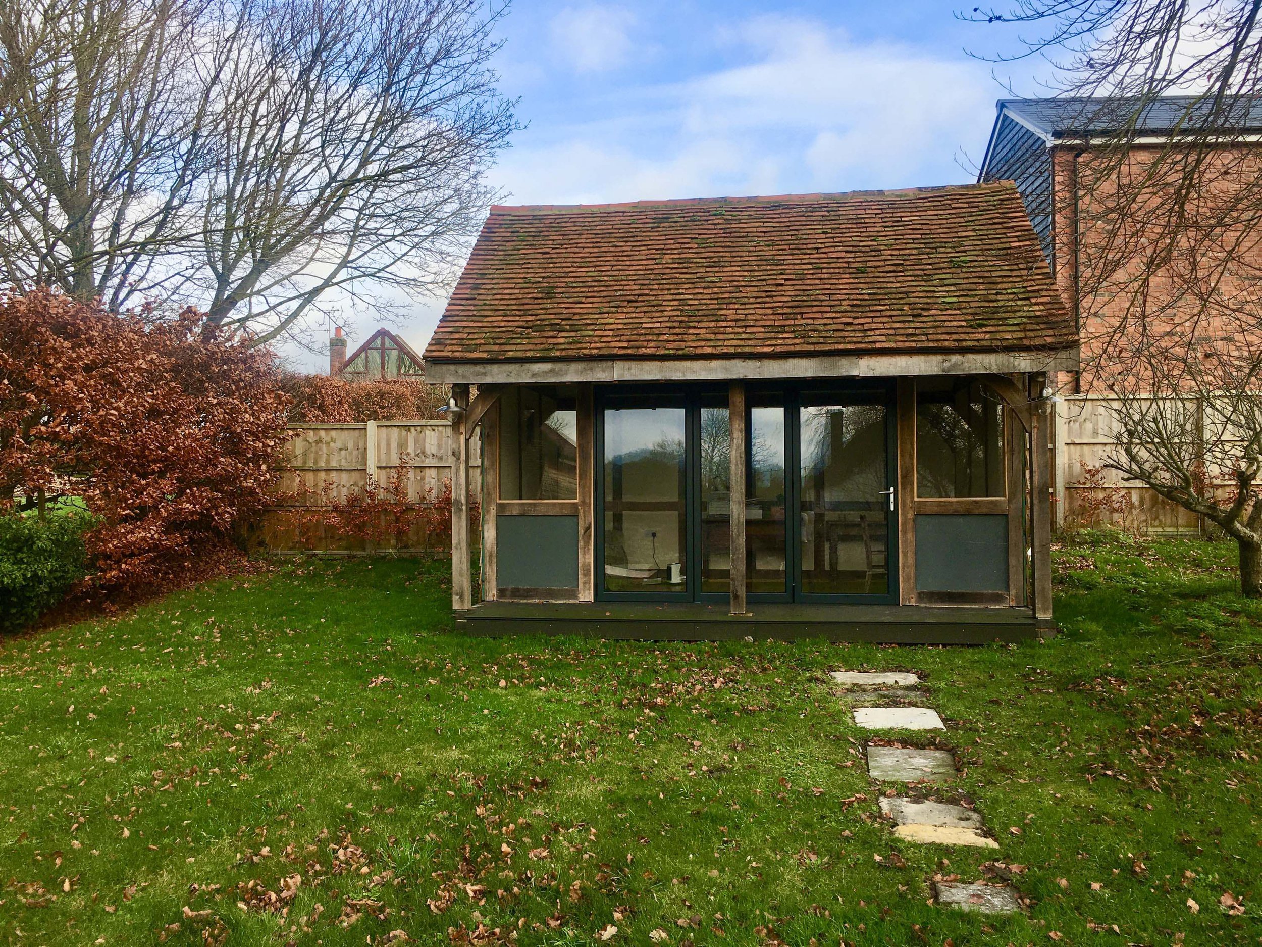 A backyard scene with a small wooden shed that has a tiled roof, a glass door, and large windows. There are leafless trees, a leafy bush, and a fenced yard with stepping stones on the grass, and a building visible in the background.