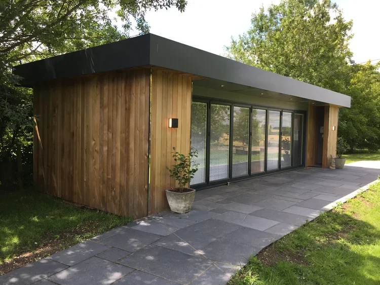 Modern single-story house with a flat roof, wooden exterior walls, large sliding glass doors, and a paved patio, surrounded by greenery and trees.