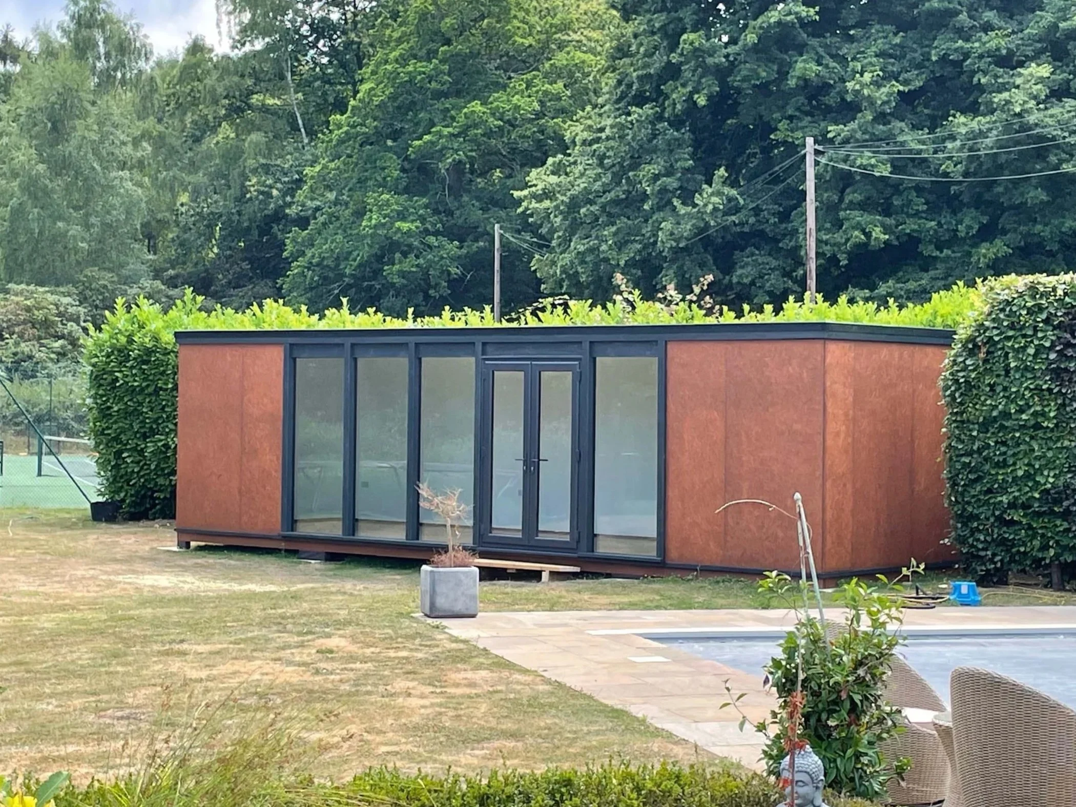A small modern building with large glass doors, set in a backyard with grass, bushes, and trees, with a tennis court visible in the background.