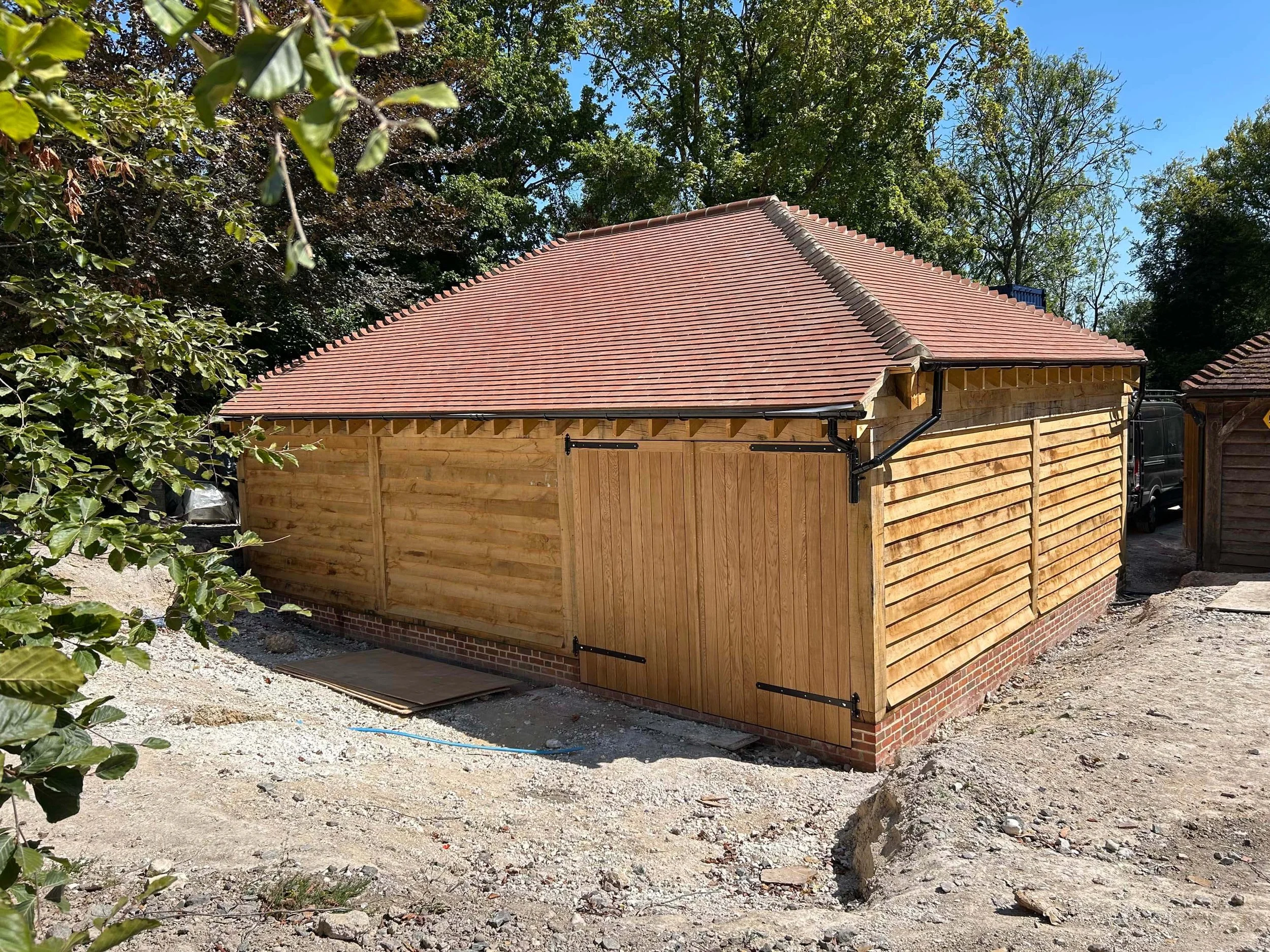 A building under construction with wooden walls and a red-tiled roof, surrounded by trees and a dirt ground.