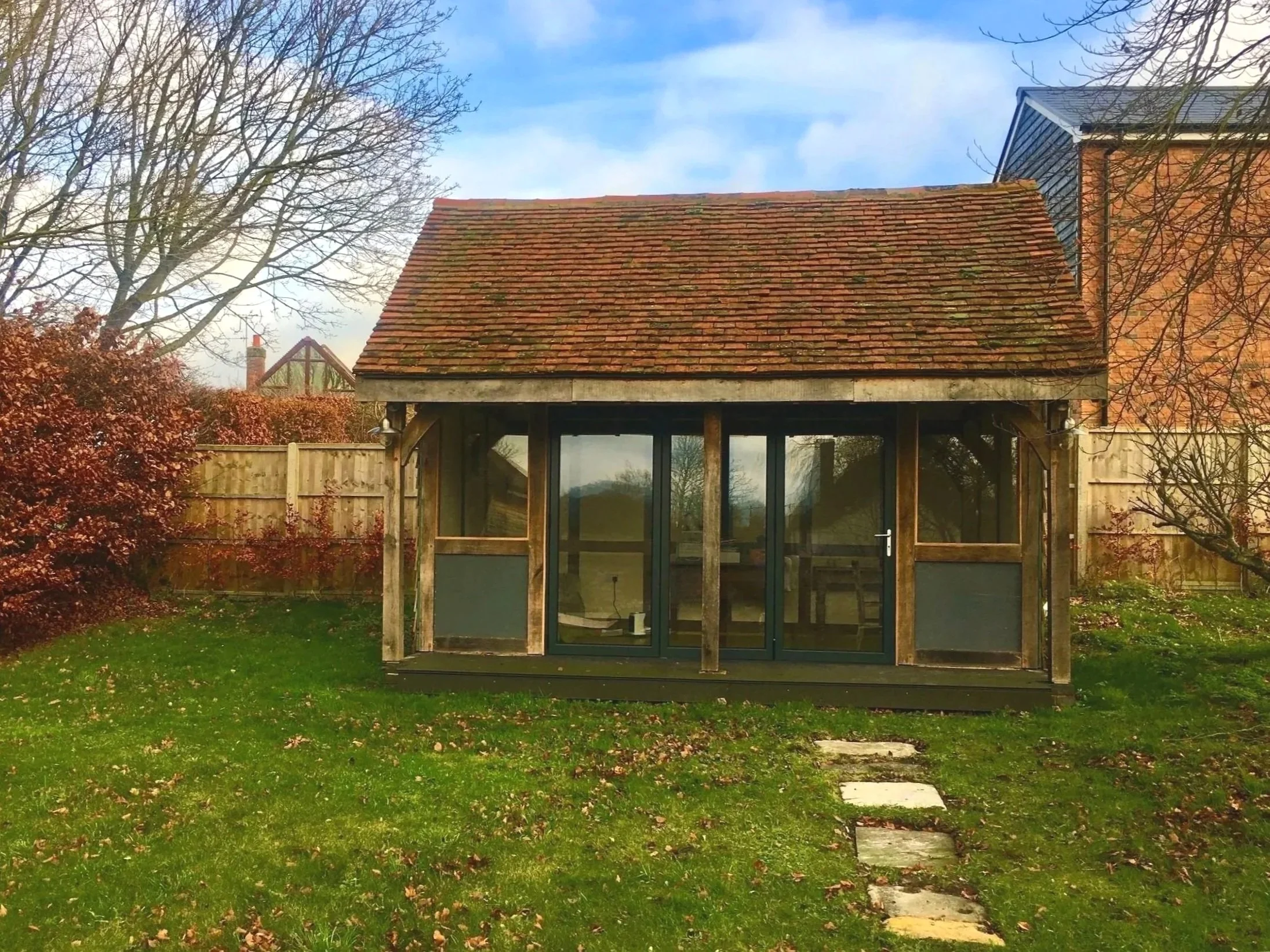 A small backyard with a newly constructed wooden garden room or shed with glass doors, surrounded by green grass, fallen leaves, trees without leaves, a bamboo fence, and a house with a blue roof in a suburban neighborhood.