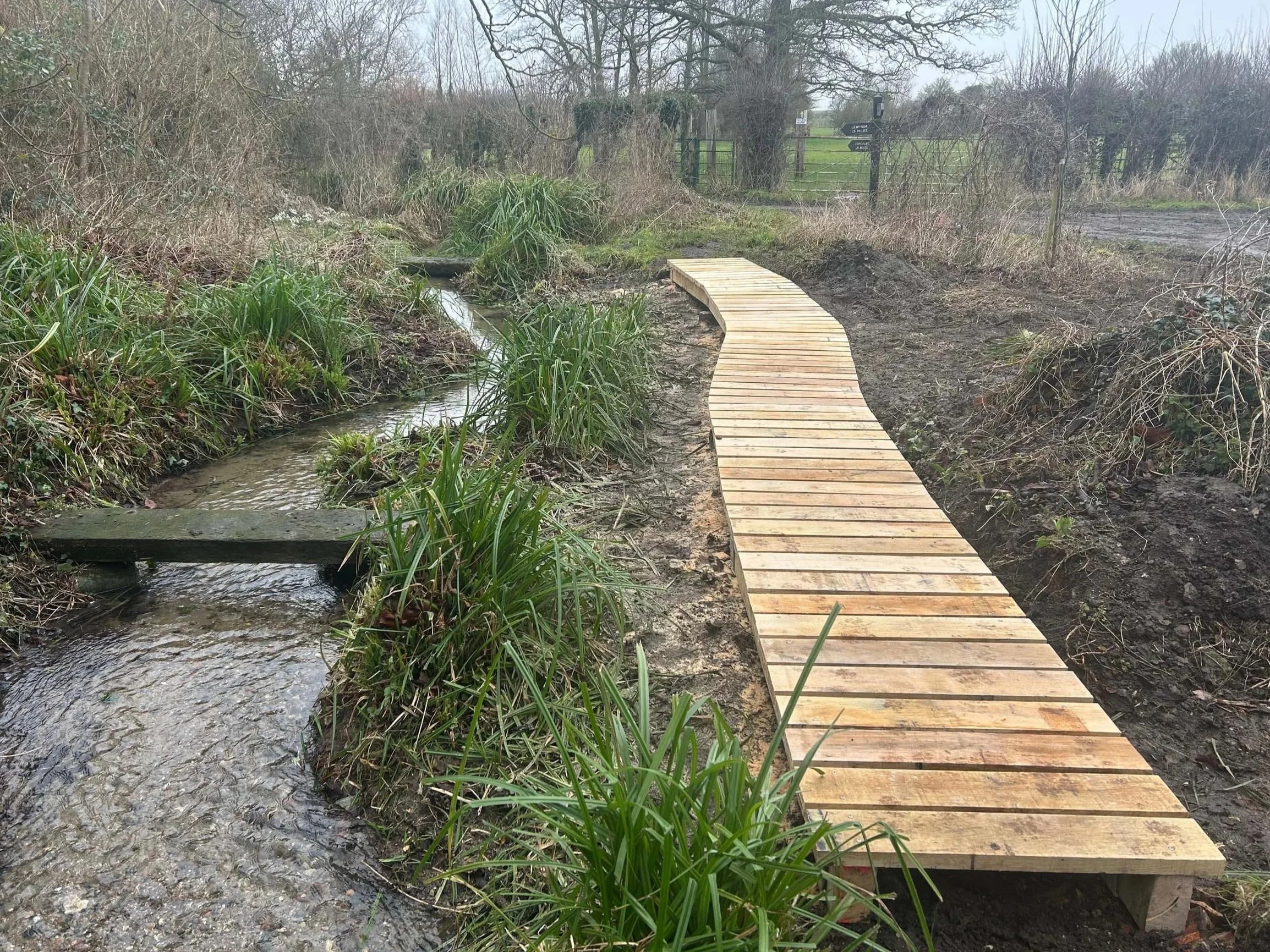 A small wooden footbridge crossing over a narrow stream in a rural area with a dirt path beyond. There are bushes and trees around, some without leaves, indicating late fall or winter. A gate and signs are visible in the distance.
