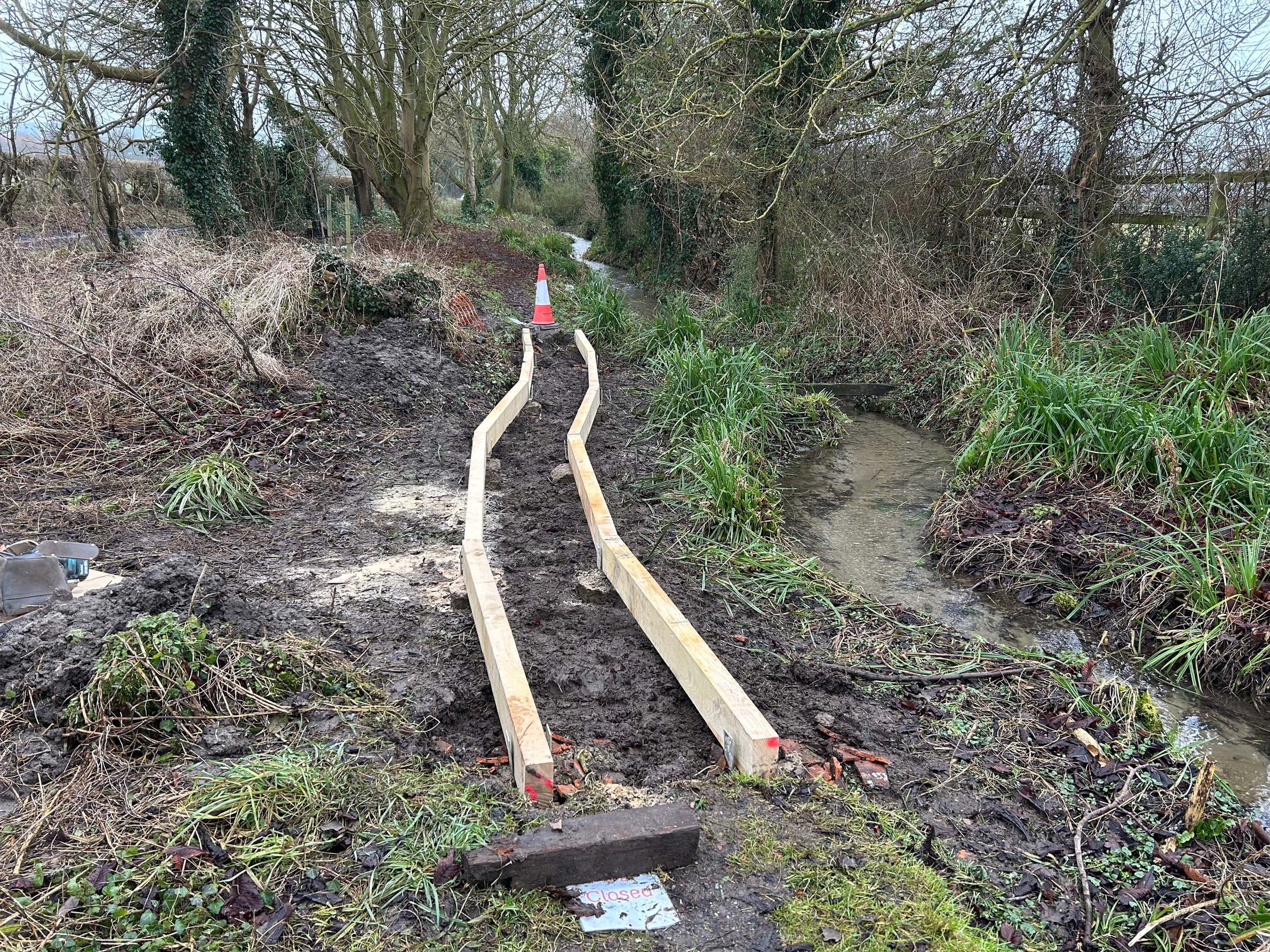 A muddy, blocked pathway with wooden rails and a small creek on the side, surrounded by leafless trees and bushes. A 'closed' sign is at the front of the path, indicating it is not accessible.